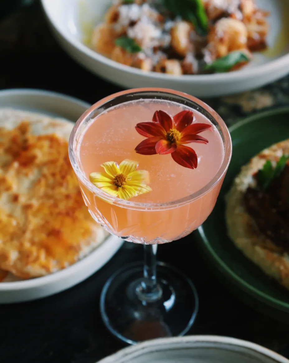 A pink cocktail with edible pink and yellow flowers floating on top, there's a pasta dish in the background and two other small plates in the background
