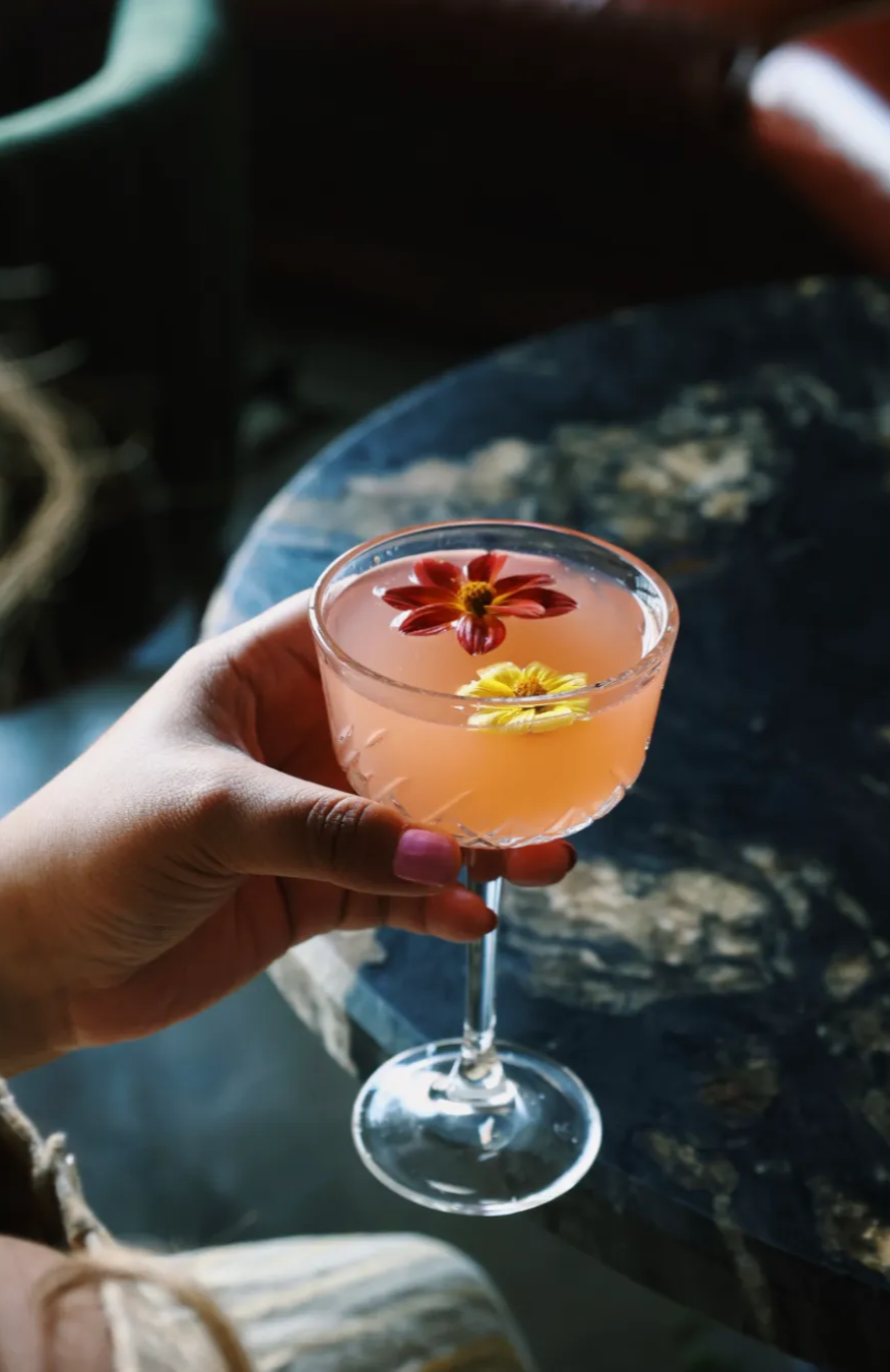 An image of a pink cocktail with a pink and yellow flower floating on top. The hand is holding the cocktail and there is a marble and black and white table in the back