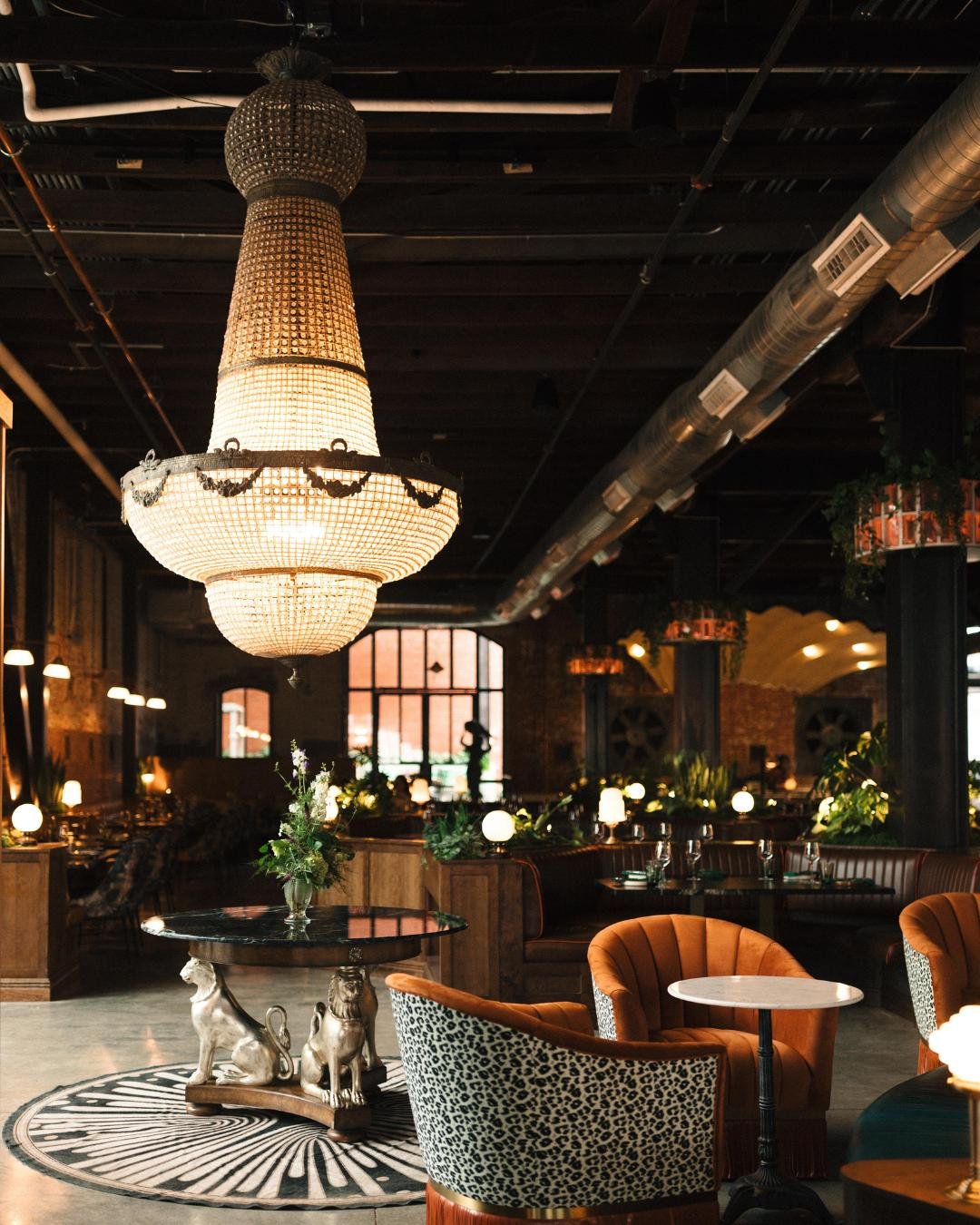 Red Herring restaurant with tables and chairs inside a large spacious old industrial building, a grand crystal chandelier hangs from the ceiling with art deco design elements including cheetah print and leather chairs. 