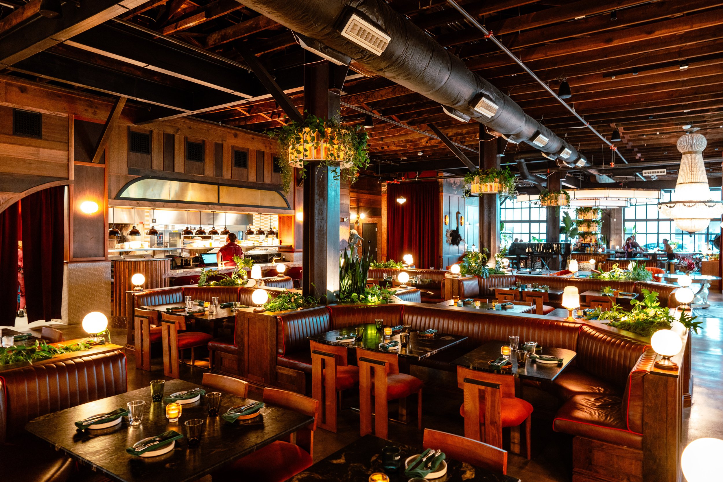 A view of the Red Herring Restaurant in a large vintage industrial style building with exposed pipes on the ceiling, warm lighting, and soft plant acccents on the columns. There is a large dining room with Leather  booths and u-shaped Tables and art 
