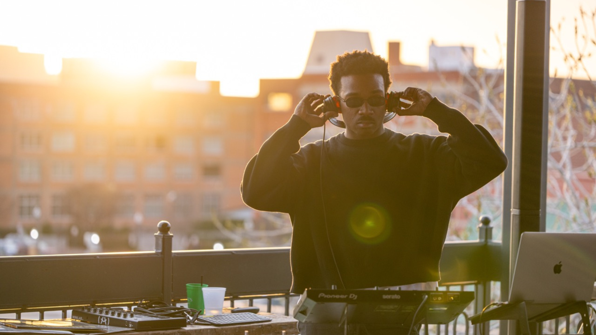 A DJ wearing sunglasses, headphones, and a black sweatshirt standing outdoors at sunset with DJ equipment and a laptop, city buildings in the background.