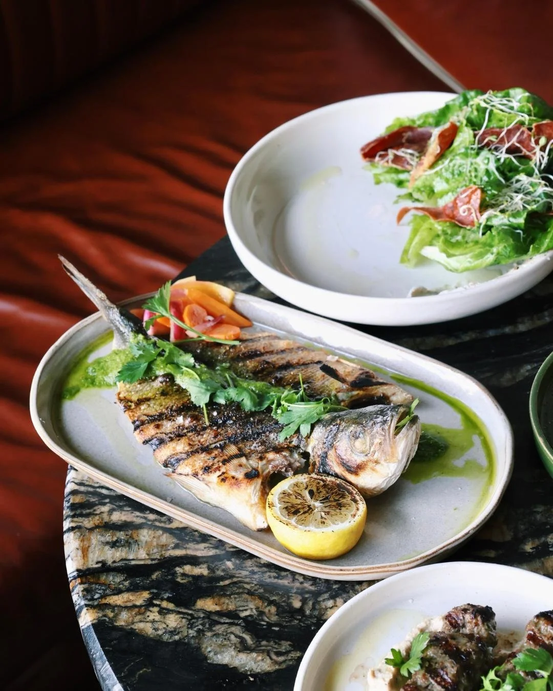 A whole food fired fish with a fresh salad and wood-fired kebabs on a black marble table.