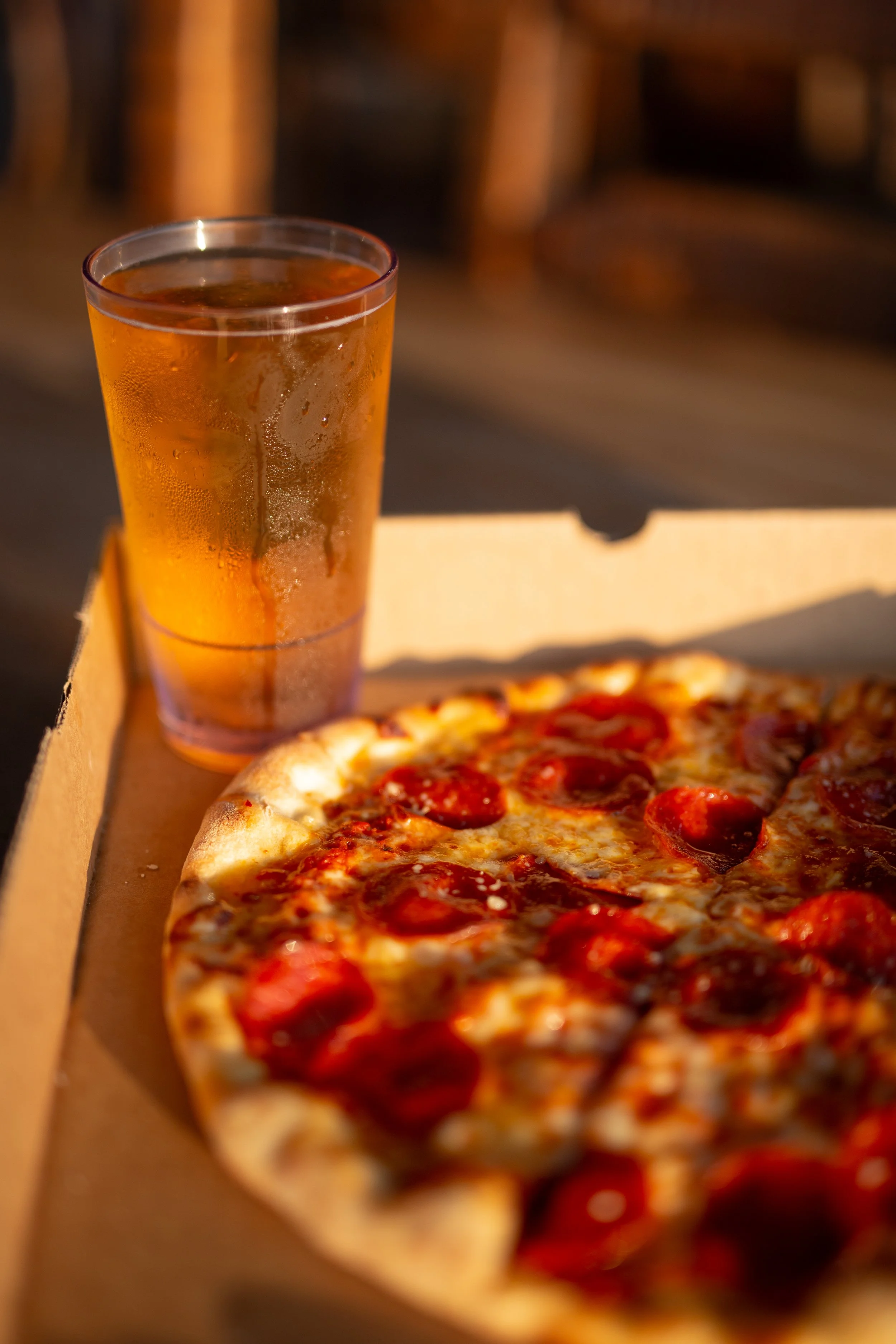 A slice of pepperoni pizza in a cardboard takeout box next to a glass of iced tea on a table.