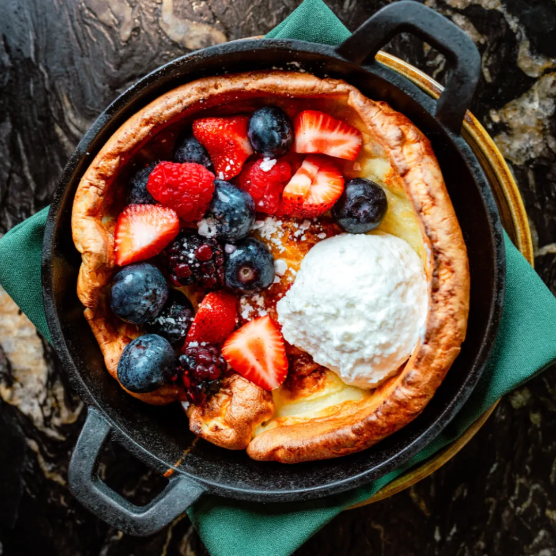 A skillet of Dutch baby pancake topped with mixed berries and a dollop of whipped cream, on a green napkin.