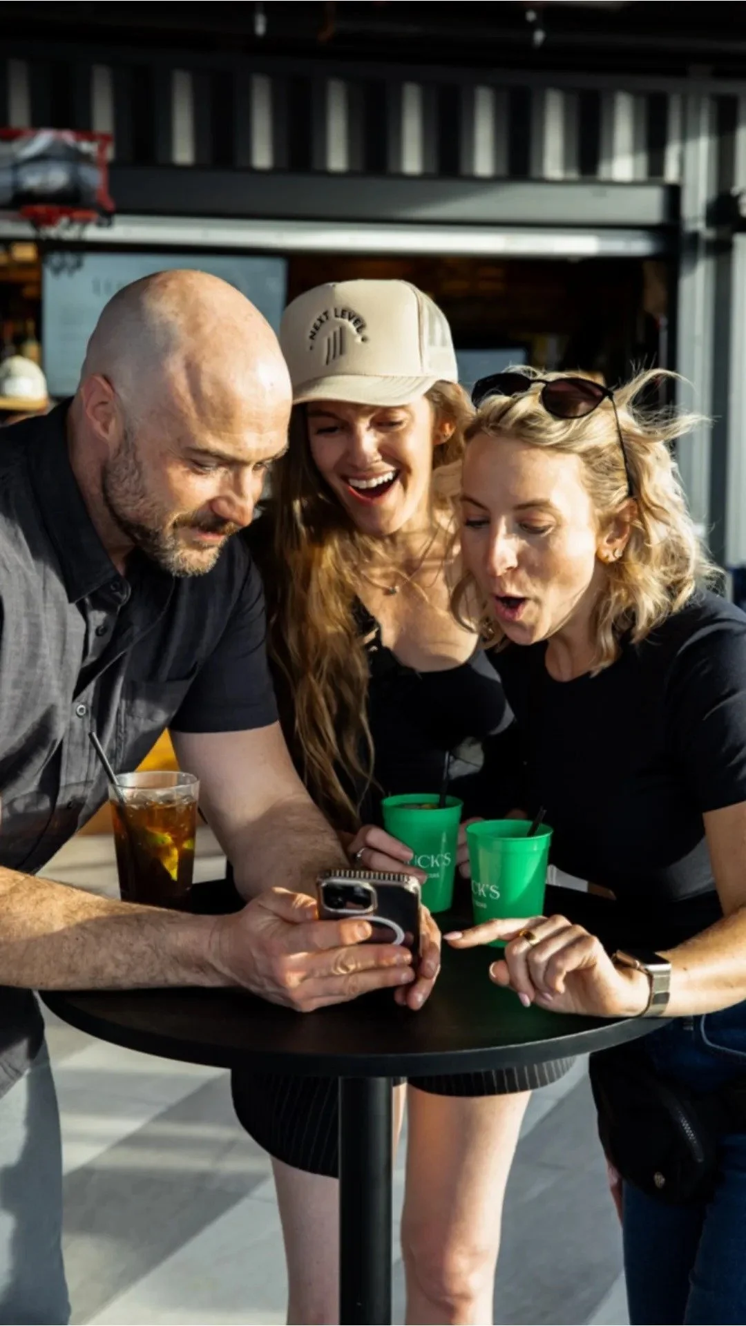 Three friends looking at a phone together at a bar, with drinks on the table, smiling and showing surprise or excitement.