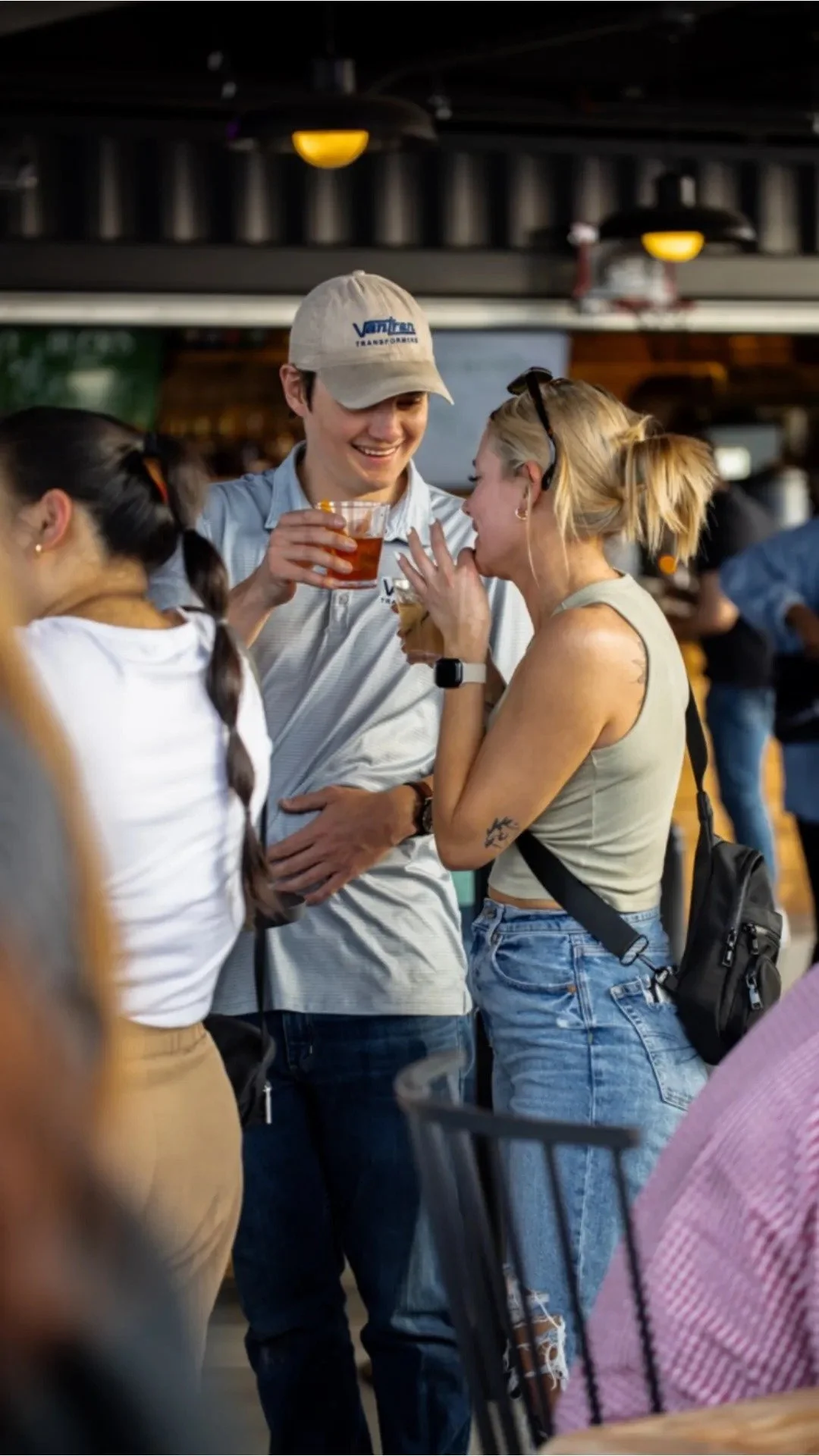 Two young women and a young man at a social gathering, smiling and holding drinks, engaged in conversation the rooftop's lively outdoor setting.