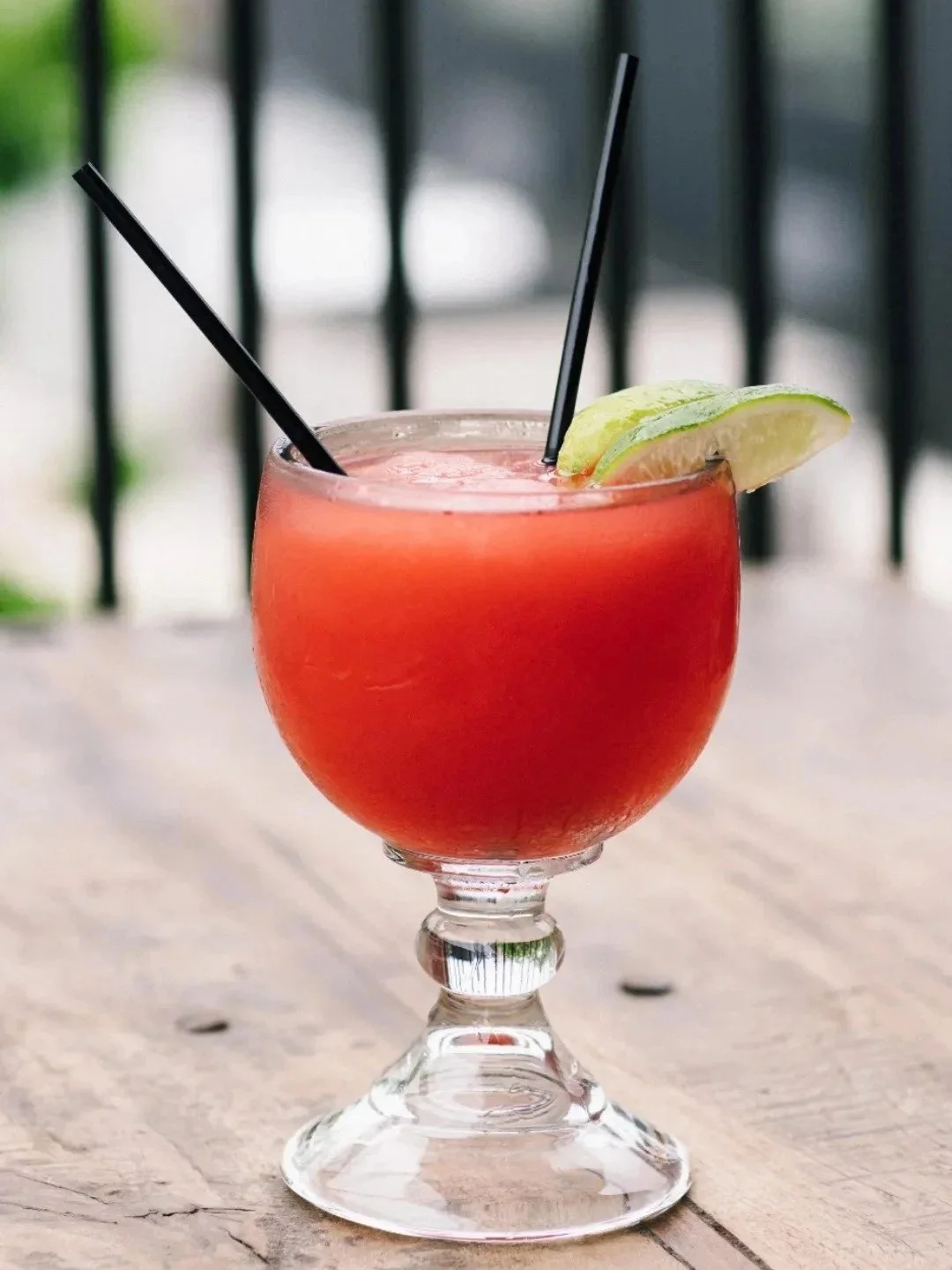 A cocktail in a rounded glass with a short stem, garnished with a lime wedge, on a wooden table with a blurred outdoor background.