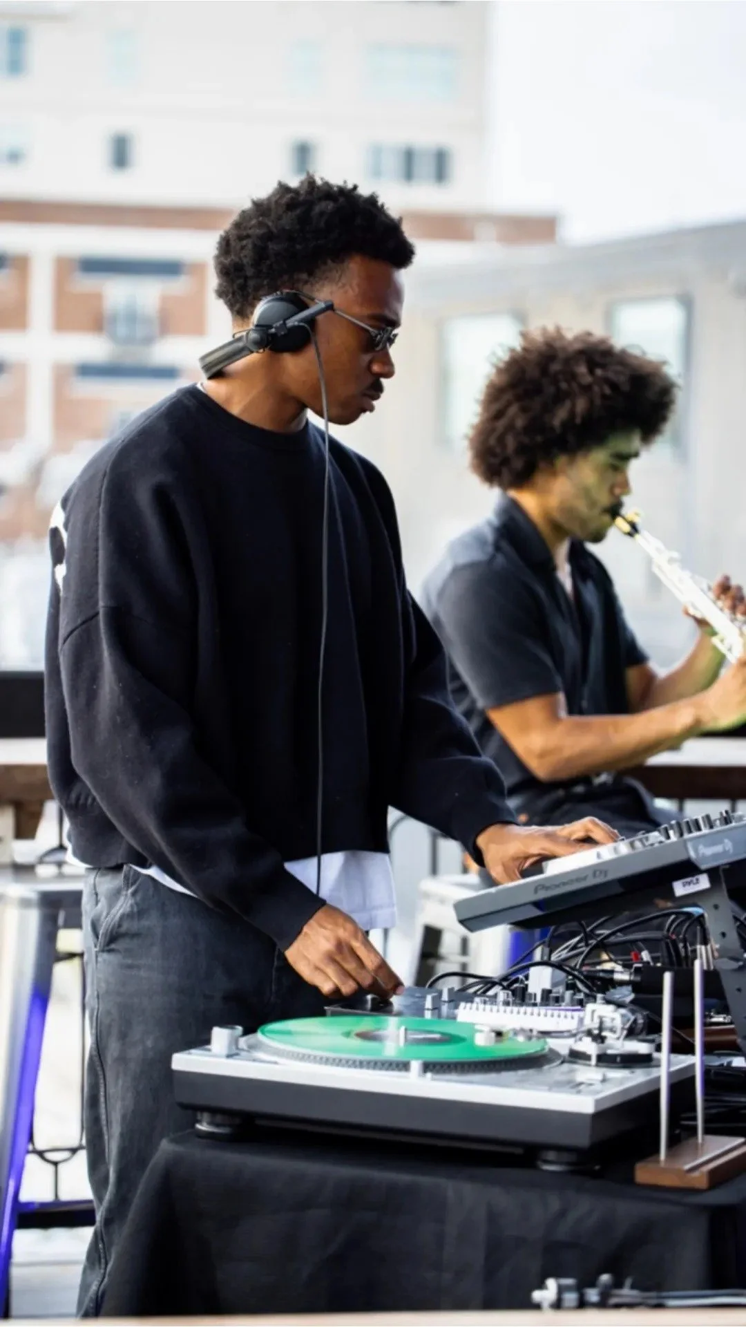 A DJ wearing sunglasses and headphones mixing music on a turntable at Lucky Buck's Rooftop Bar, with a man in the background playing an instrument.