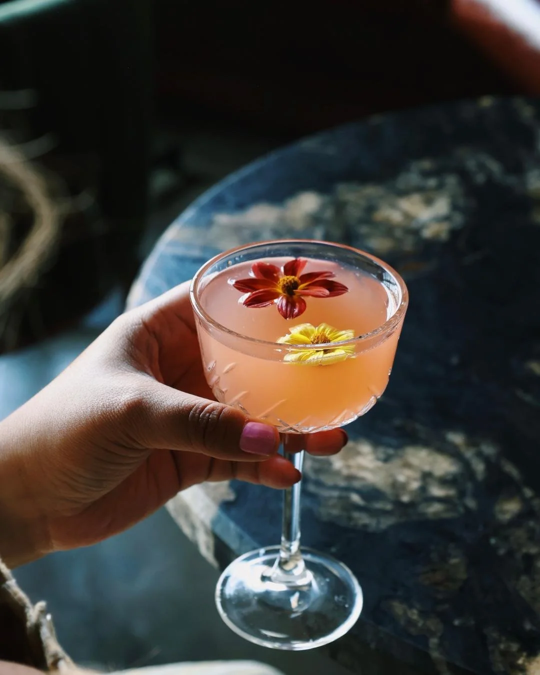 A hand holding a pink cocktail with floating edible flowers on top of it