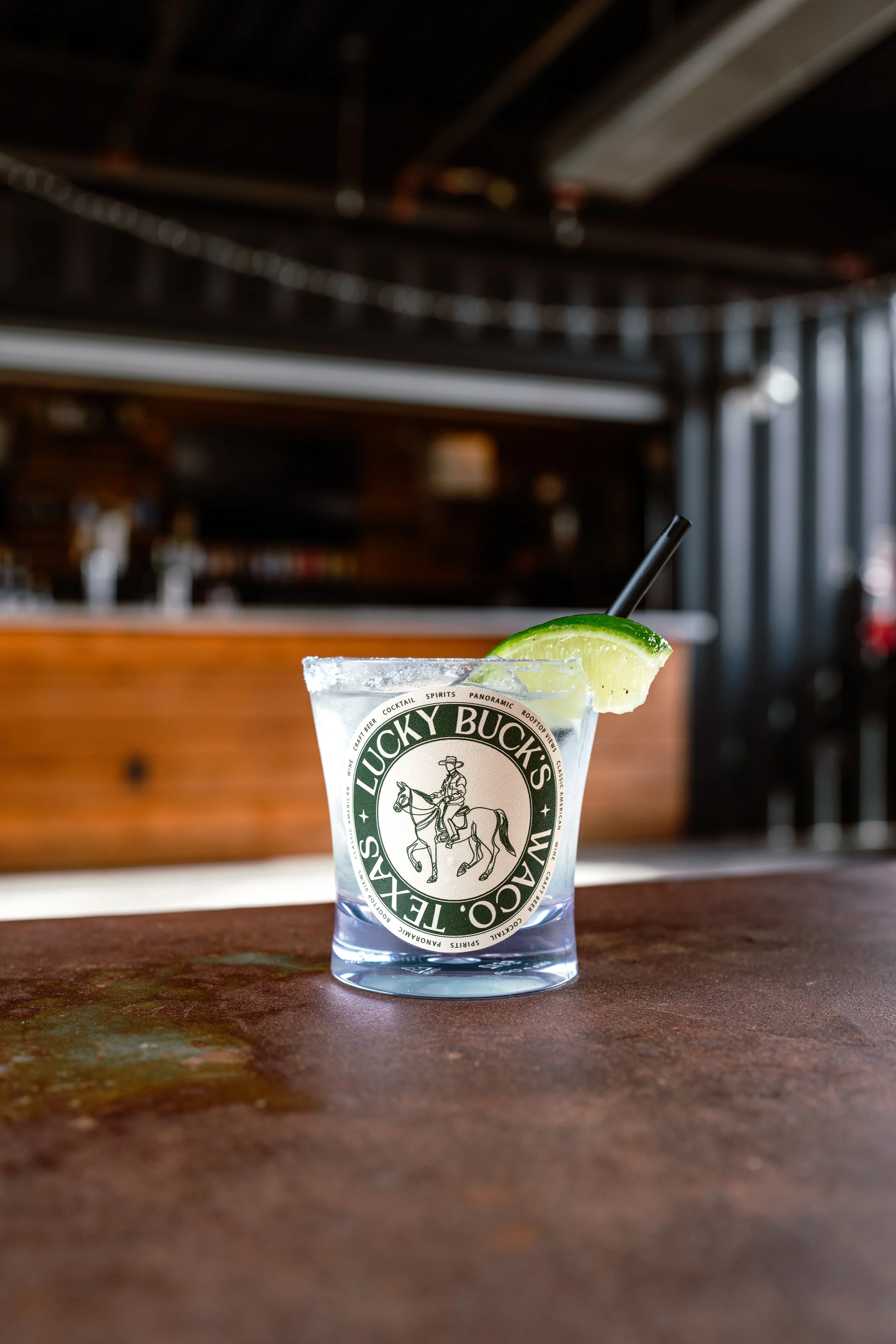 A glass of clear cocktail with lime garnish and a straw, placed on a wooden bar counter, with a blurred bar background.