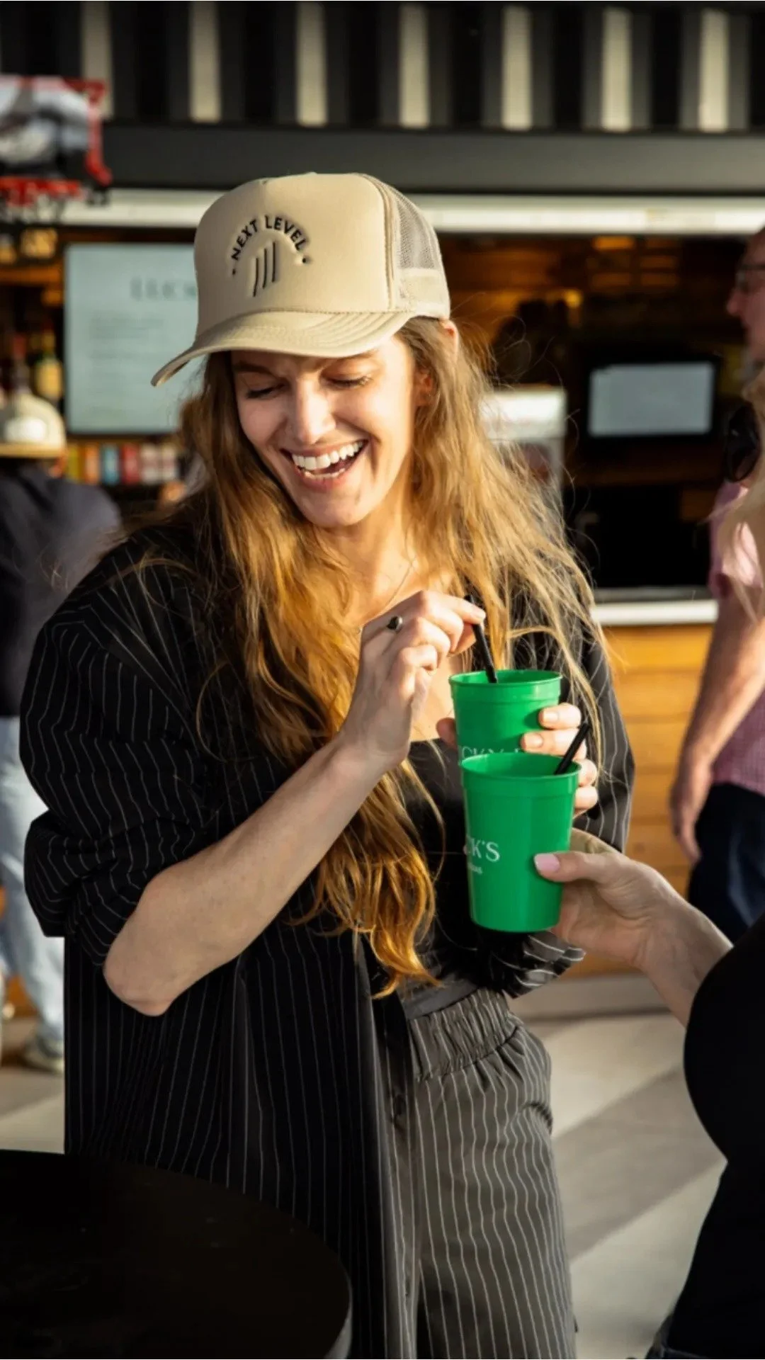 Woman with long red hair smiling and holding two green Lucky Buck's cups with black straws, wearing a beige baseball cap and a black shirt.
