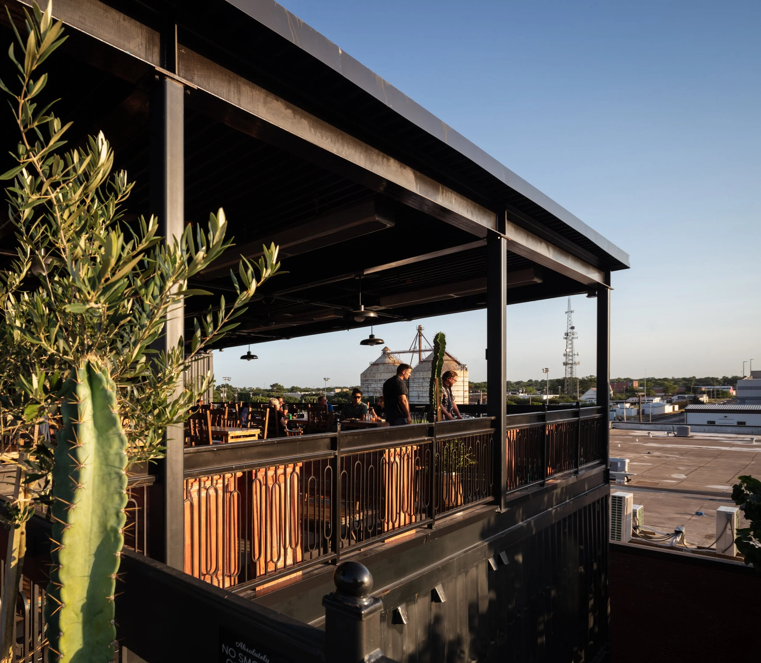 People on a rooftop terrace in the late afternoon sun, with potted plants and cityscape in the background.