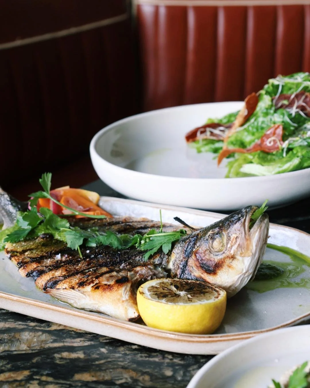 A whole cooked fish with a charred lemon, the fish is wood-fired, there is a plate of salad in the background
