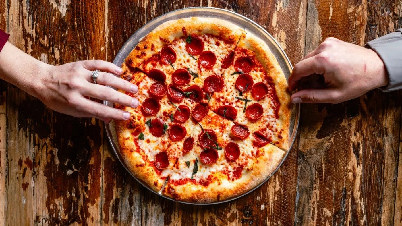 A pizza with pepperoni slices on a wooden table, with two hands reaching toward it.