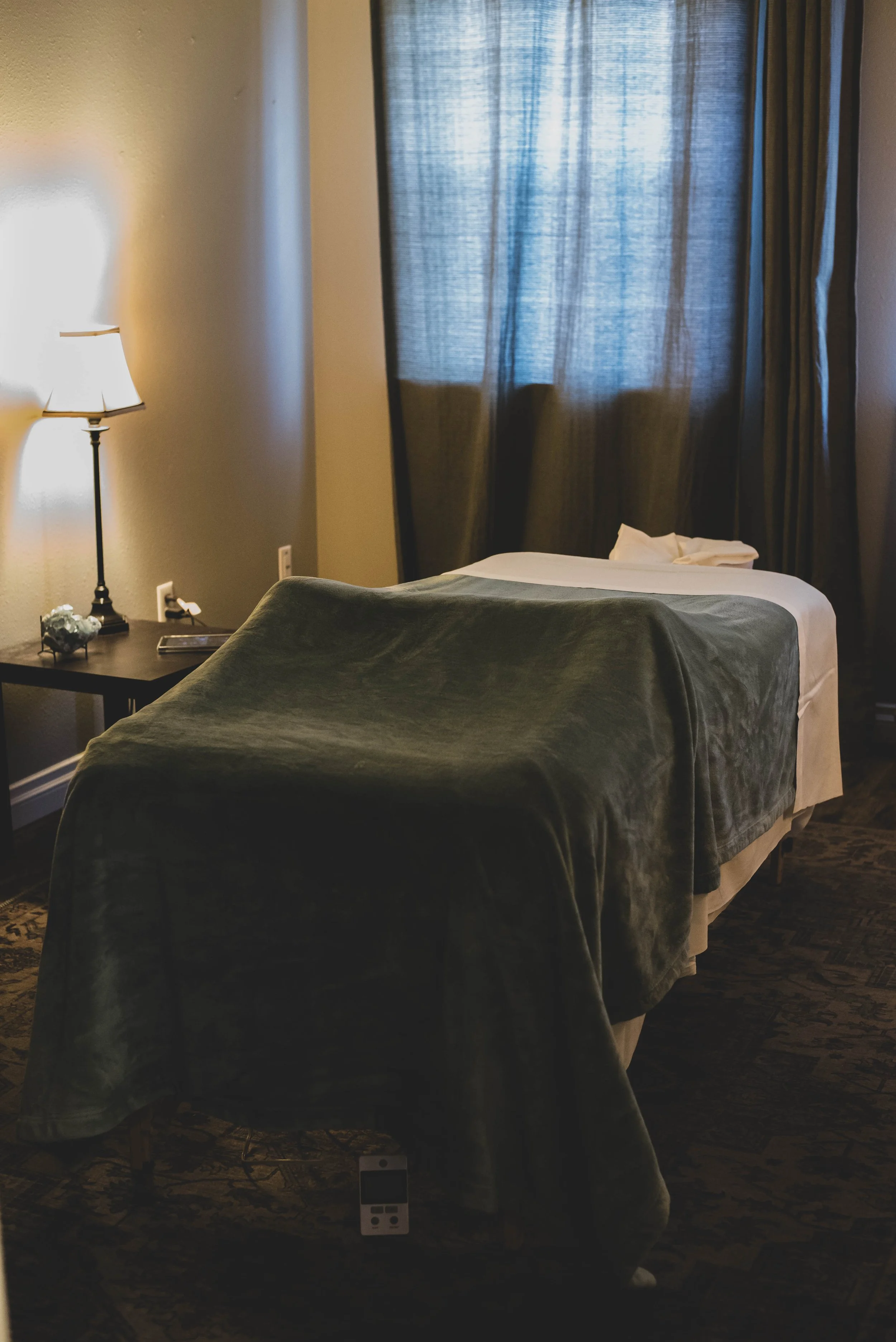 A hotel room with a bed covered in a dark green blanket, a bedside table with a lamp, and a window with dark curtains that are partially open. There is a towel on the bed and a remote control on the floor.