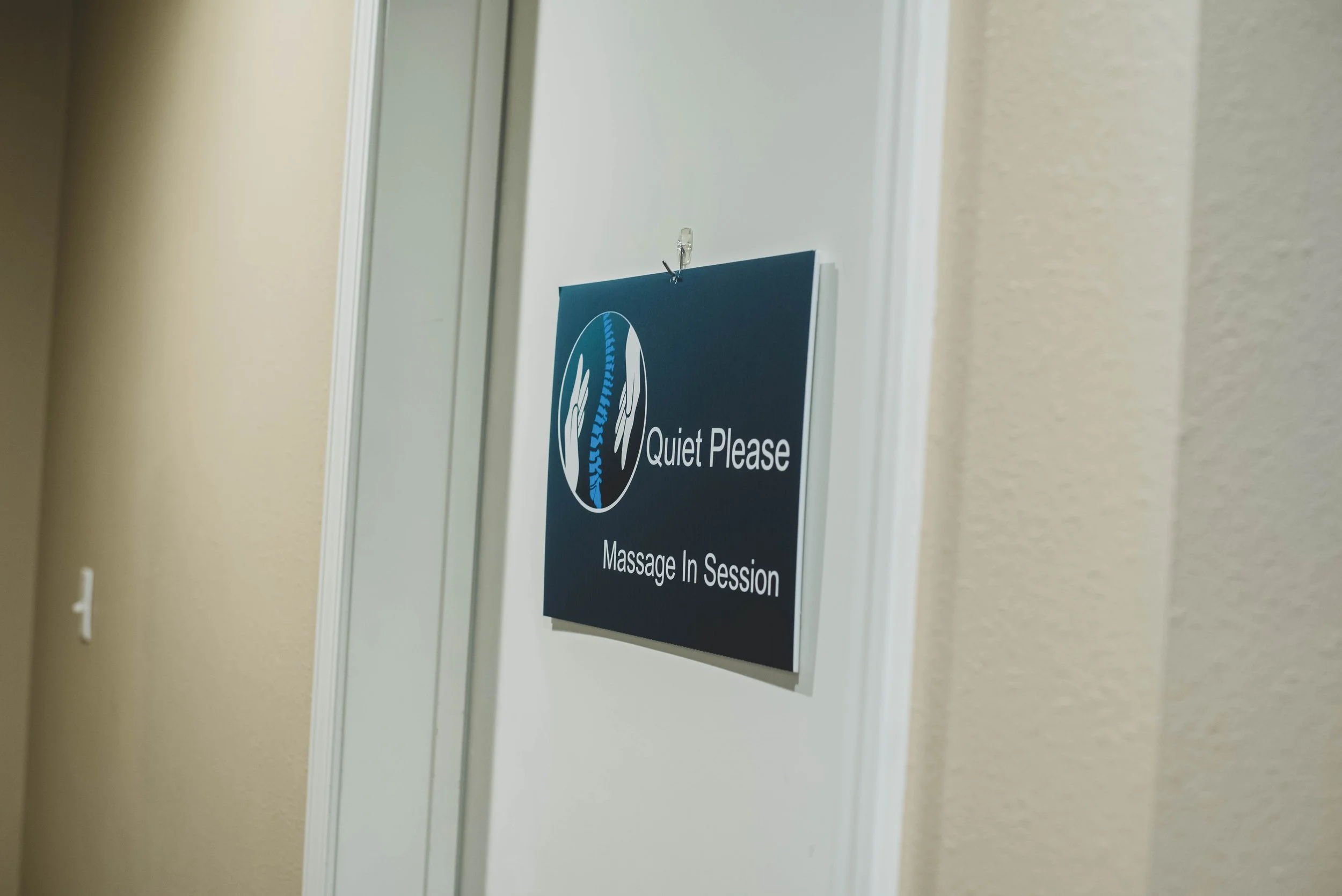 A blue sign on a white wall indicating a massage in session with a logo of two hands and a spine, and the text 'Quiet Please.'