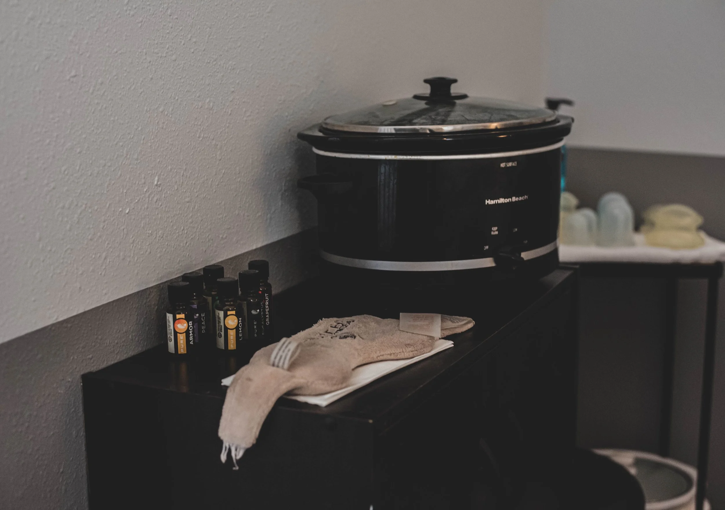 A black tabletop with essential oils, a dish towel, and a white cloth, with a Hamilton Beach slow cooker on a gray wall behind.