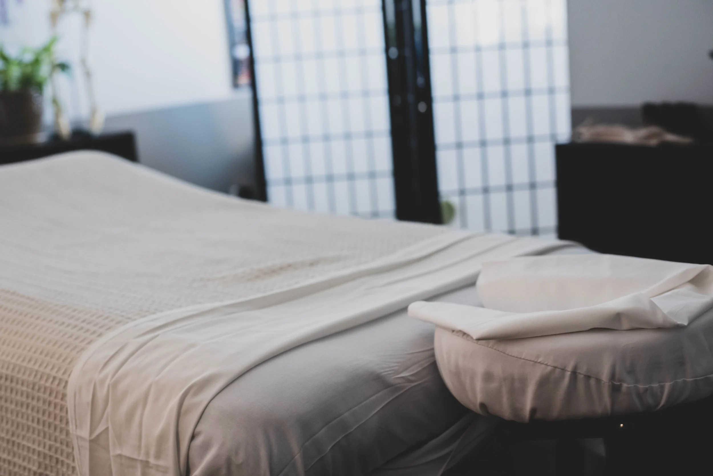 Close-up of a neatly made bed with cream-colored sheets and textured blanket, with a blurred background of a window with bars and some household items.
