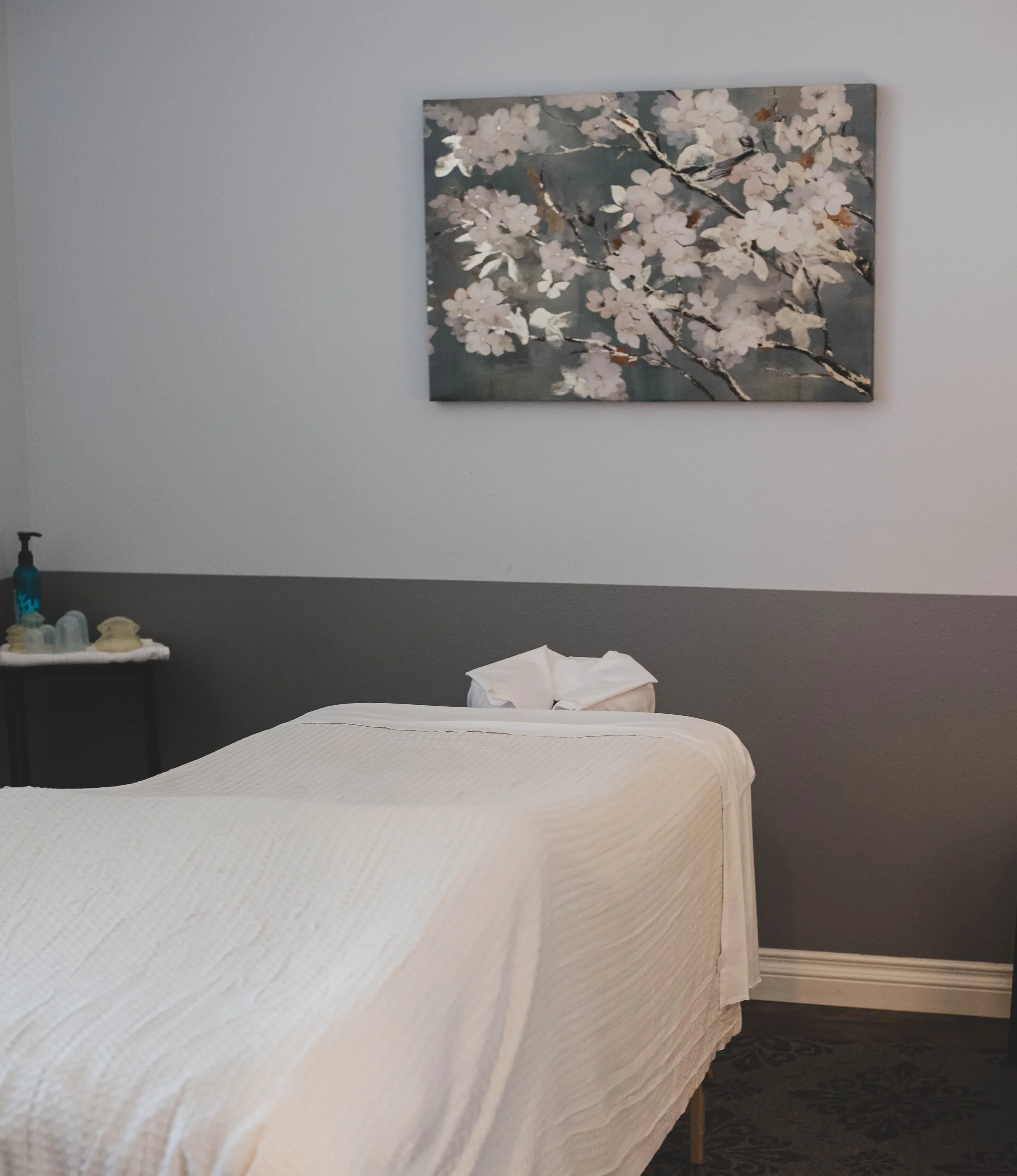 Hotel room with a bed covered in a white textured blanket, a tissue box, and a side table with toiletries. A large painting of cherry blossoms on a dark background hangs above the bed.