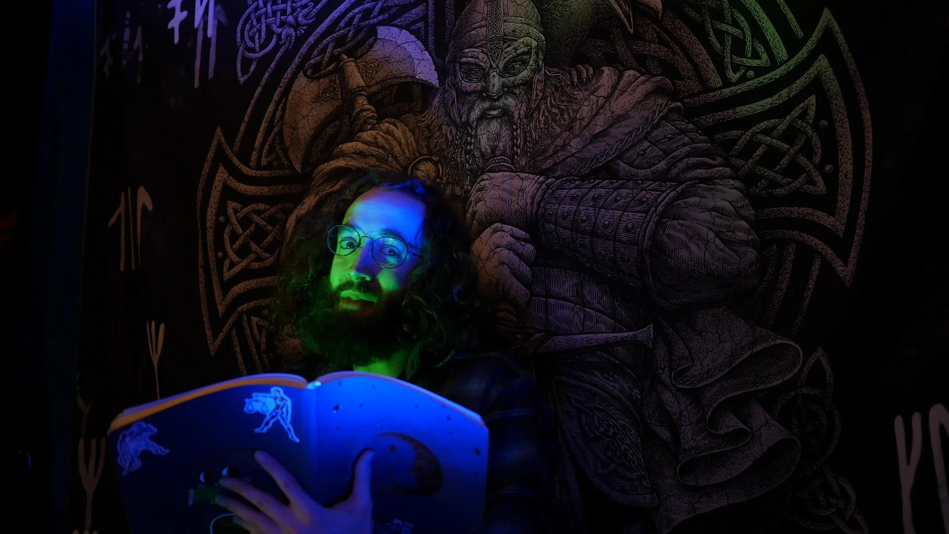A person with long curly hair, glasses, and a beard holds an open book, illuminated by green, blue, and purple lights. Behind them is a large chalk drawing of a Viking warrior with a helmet, armor, and beard, holding a sword and shield, on a dark background.
