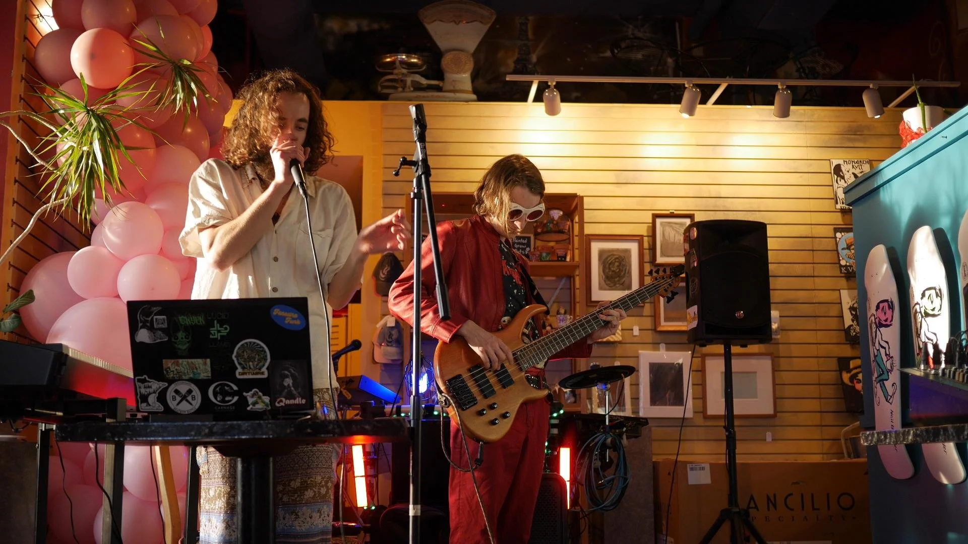 Two musicians performing with instruments on a colorful, cozy stage decorated with pink balloons, artwork, and framed pictures.