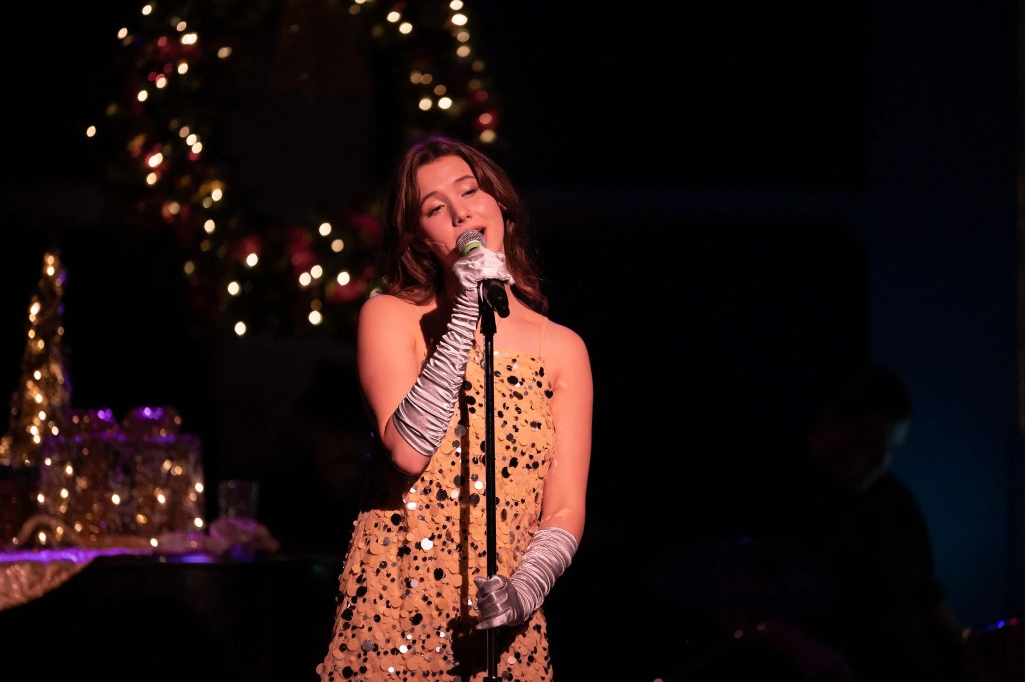A young woman singing into a microphone on stage, wearing a sleeveless, gold dress with large sequins and long silver gloves, with decorated Christmas tree in the background.