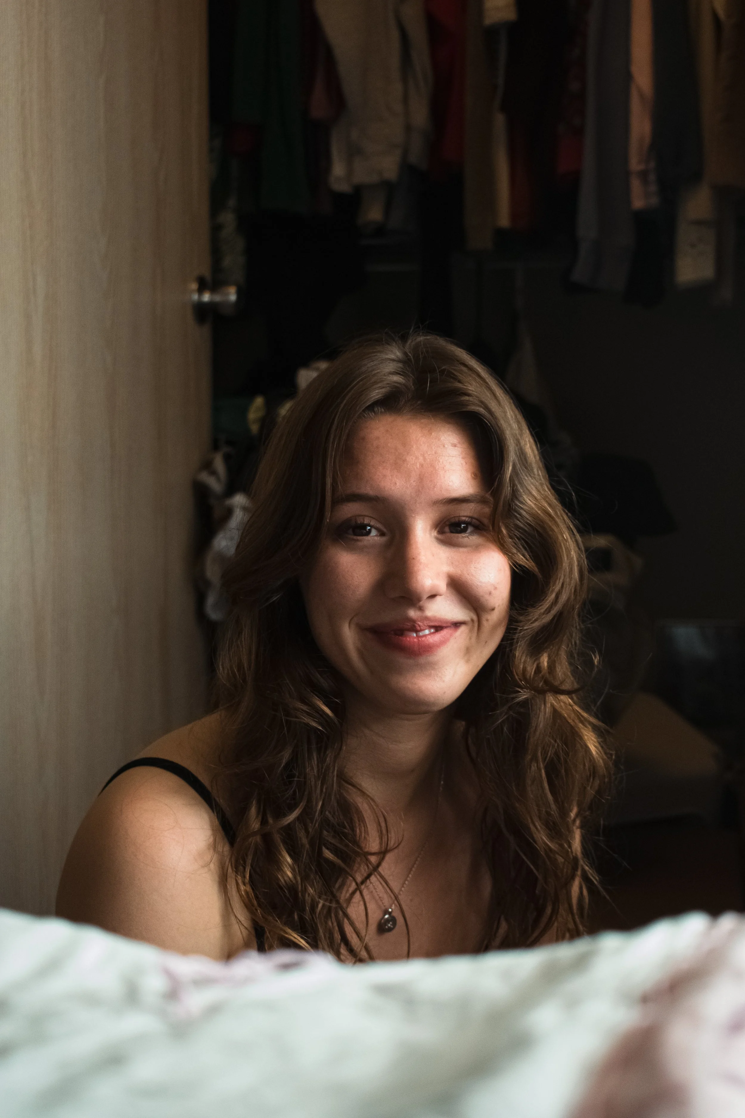 A young woman with wavy brown hair and freckles, smiling at the camera, in a room with an open closet in the background.