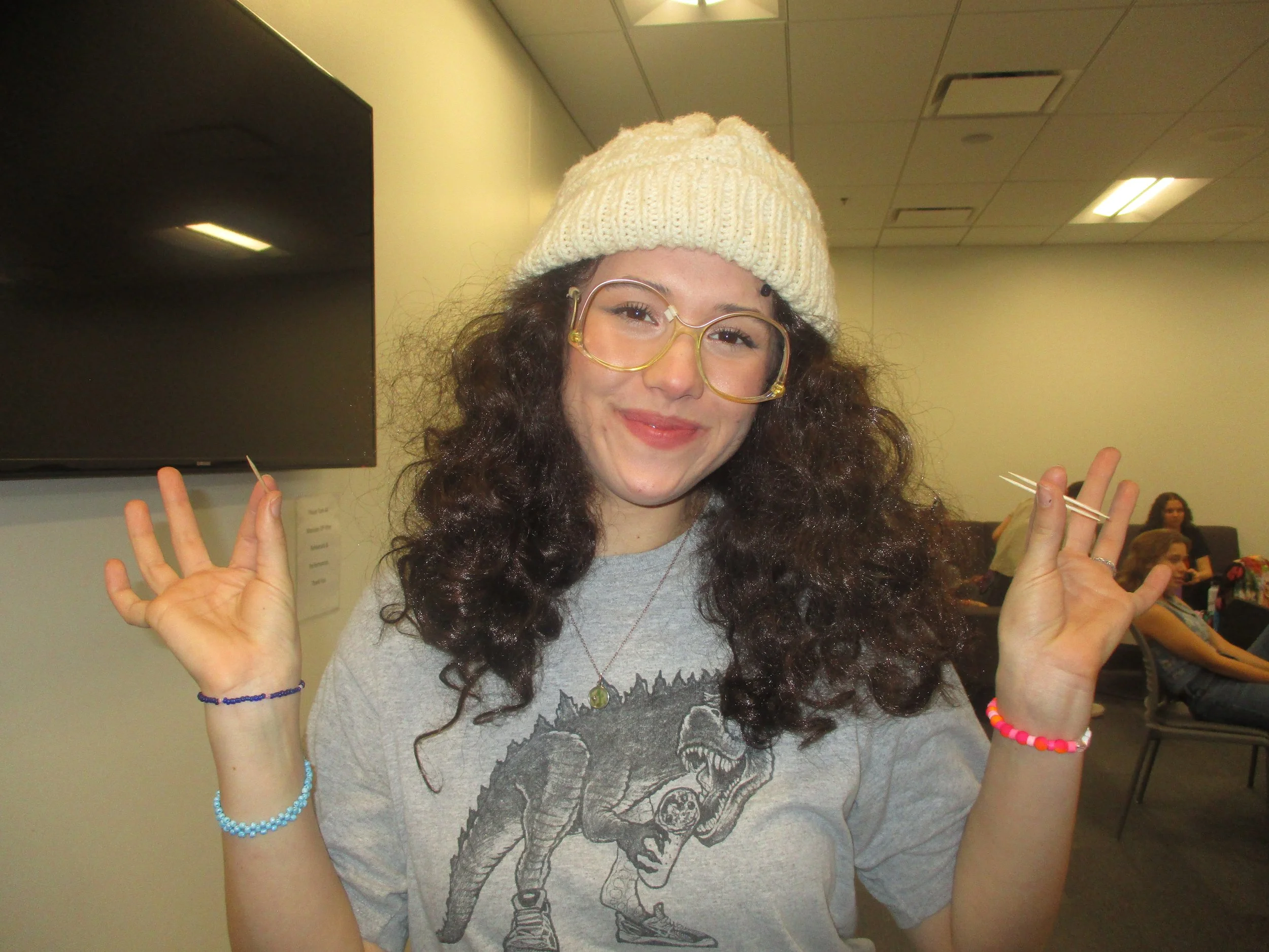 A young woman with curly dark hair wearing a white knit beanie, yellow glasses, a gray graphic T-shirt, and colorful bracelets holds two toothpicks in her hands and smiles at the camera. She is in a room with a black television and several people sitting in the background.