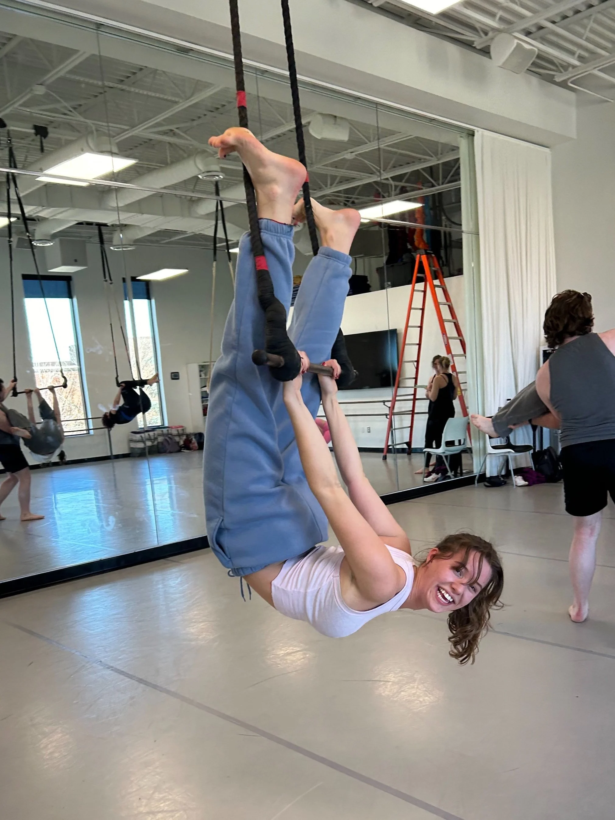 Young woman smiling and hanging upside down by her knees from aerial silks in a dance or fitness studio, with a mirror and other people practicing in the background.