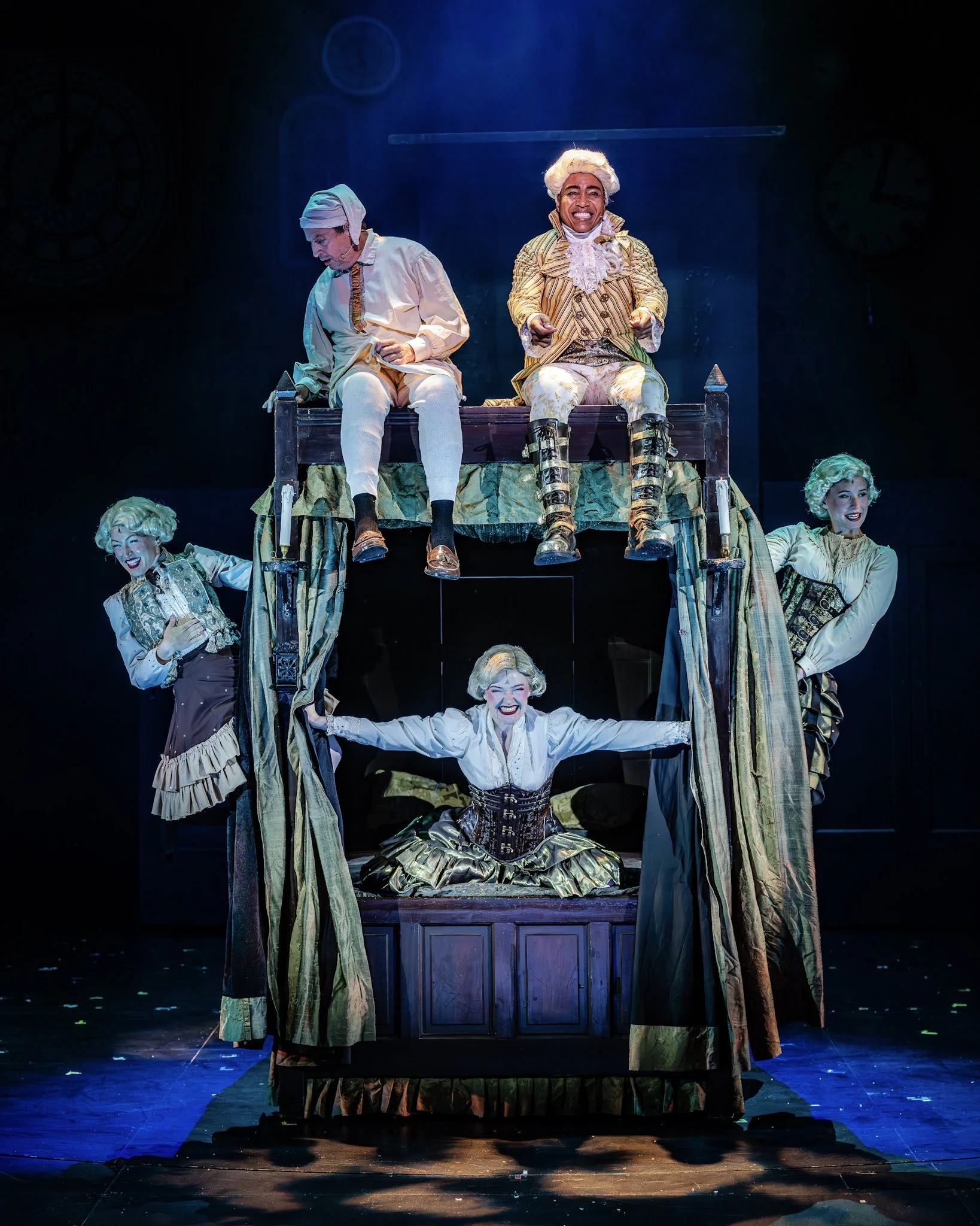 Theater scene with actors in 18th-century costumes on a multi-level platform with drapes, smiling and posing dramatically.