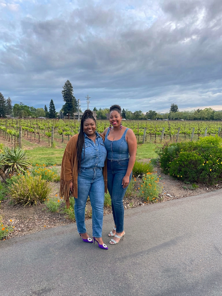 Two women standing outside in front of a vineyard, dressed in denim jeans and heels, smiling at the camera.