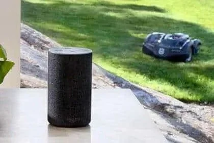 A black smart speaker on a table in the foreground, with a robotic lawn mower on the grass in the background.