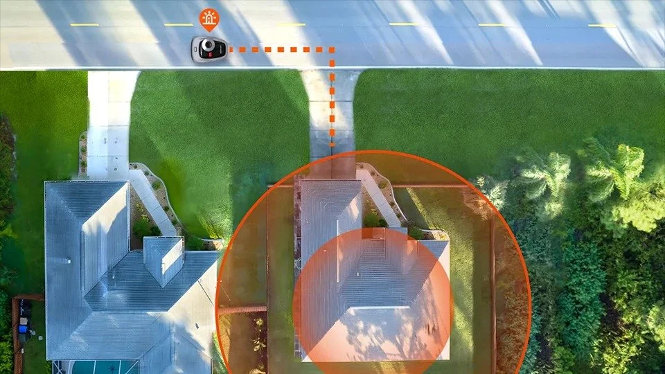 An aerial view of a house with a driveway, a lawn, and a car at the top. The image shows a Wi-Fi signal range overlay around the house and driveway, with a spotter camera and an orange alert icon on the road.