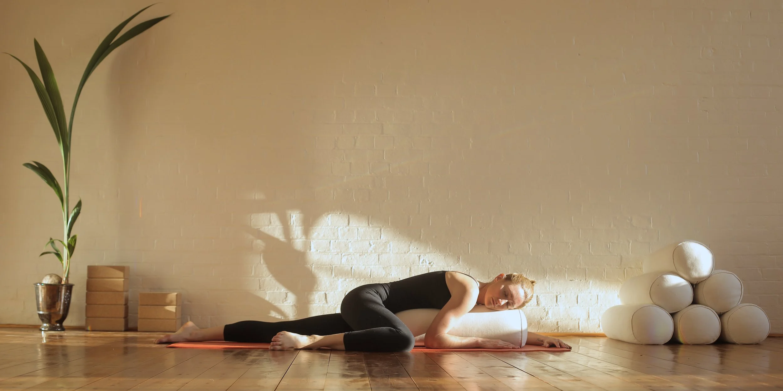 A woman on a yoga mat uses a foam roller under her neck during a stretch in a bright, minimalist room with a white brick wall, hardwood floor, houseplant, and stacked yoga blocks.
