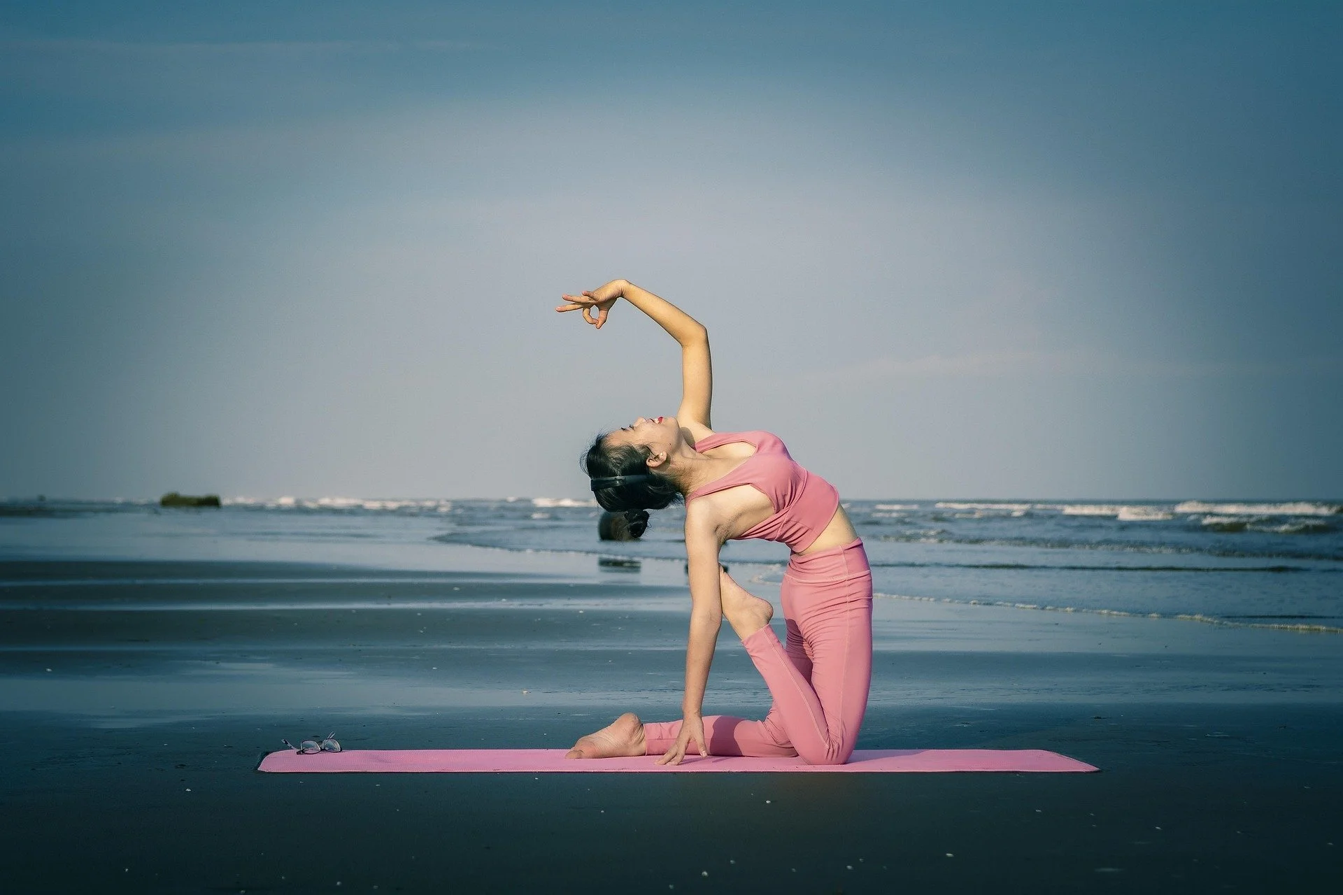 Woman practicing yoga on a pink mat on the beach, performing a backbend pose with an ocean view in the background.