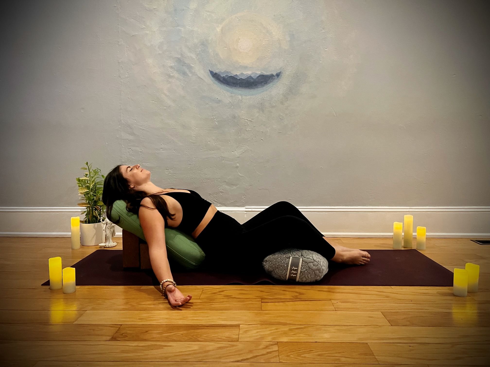 A woman practicing yoga or relaxation in a room with candles, a plant, and a large mural of a moon on the wall.