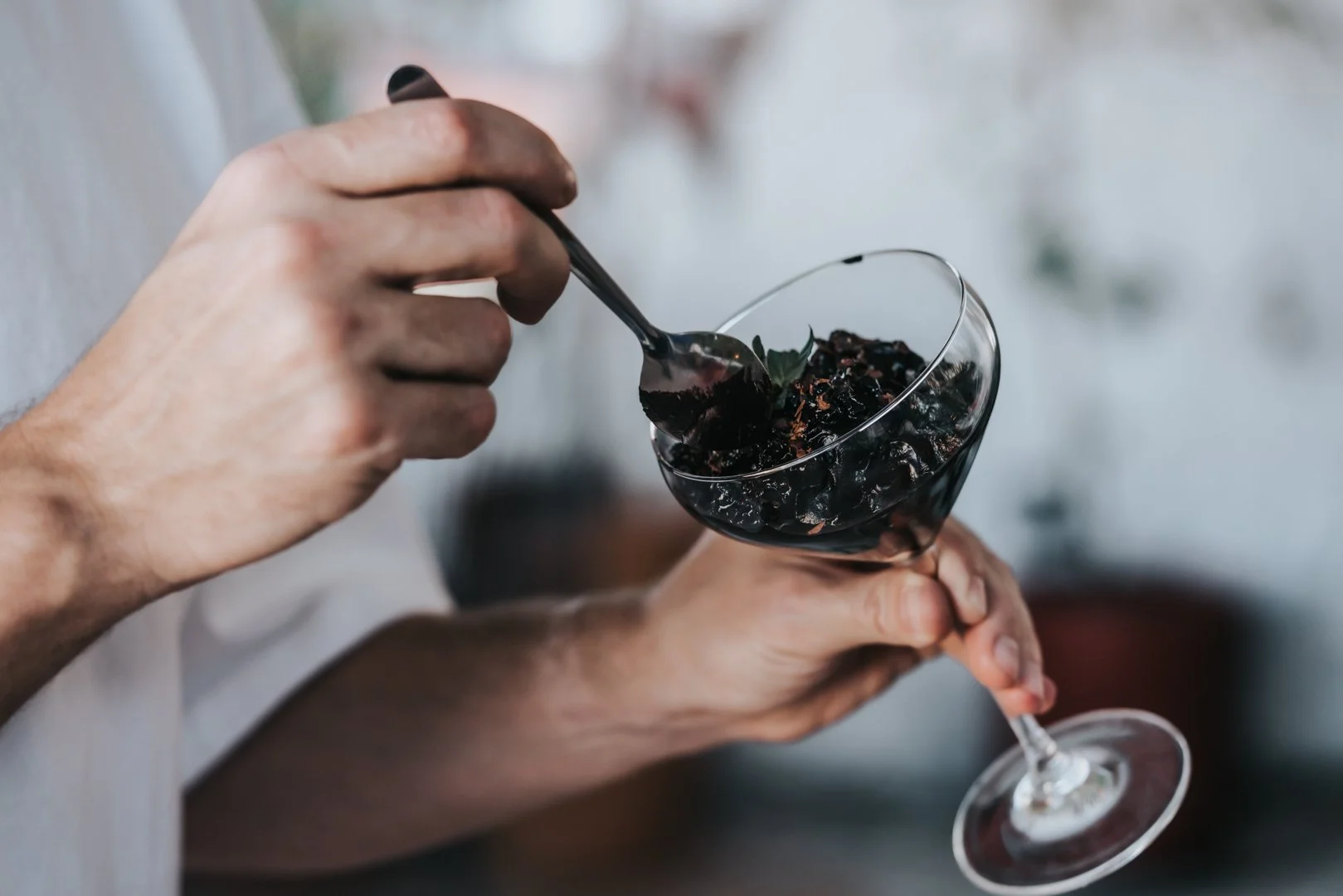 Una mano sostiene el tallo de una copa de cóctel llena de mousse de chocolate y aguacate, decorada con una ramita de menta. Otra mano mete una cuchara, lista para tomar un bocado.