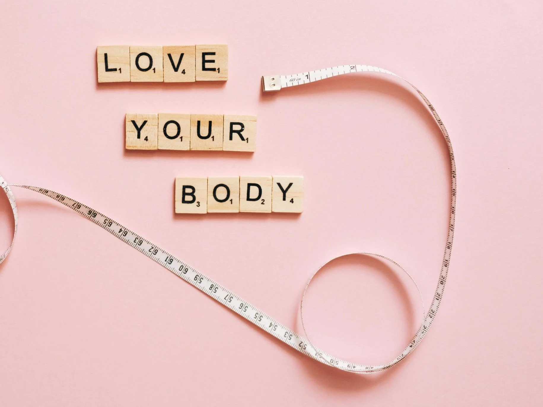 Scrabble tiles spelling 'LOVE YOUR BODY' next to a measuring tape on a pink background.
