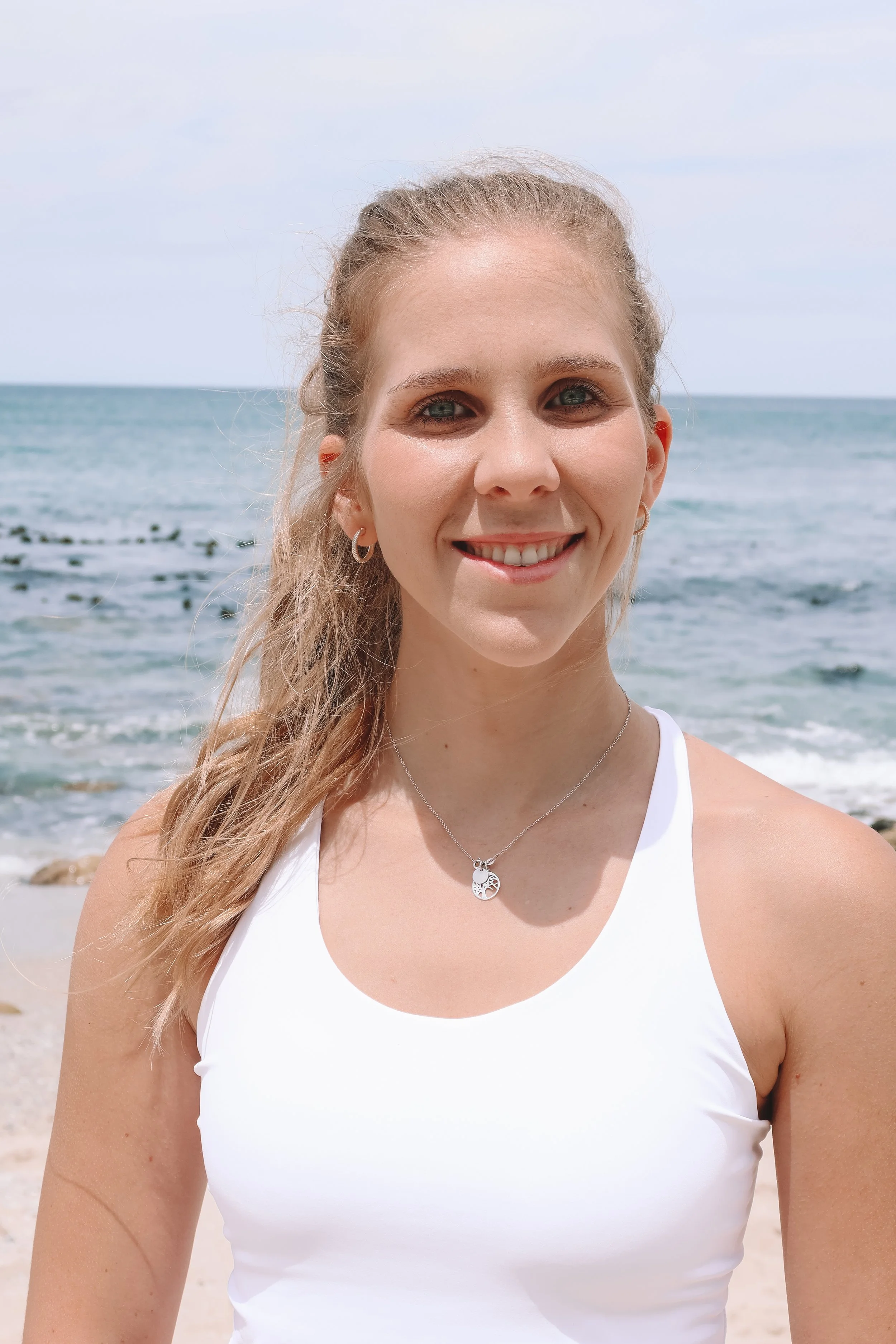 Health coach in a white exercise top standing on a beach with ocean waves in the background, wanting to help you reach your exercise goals.