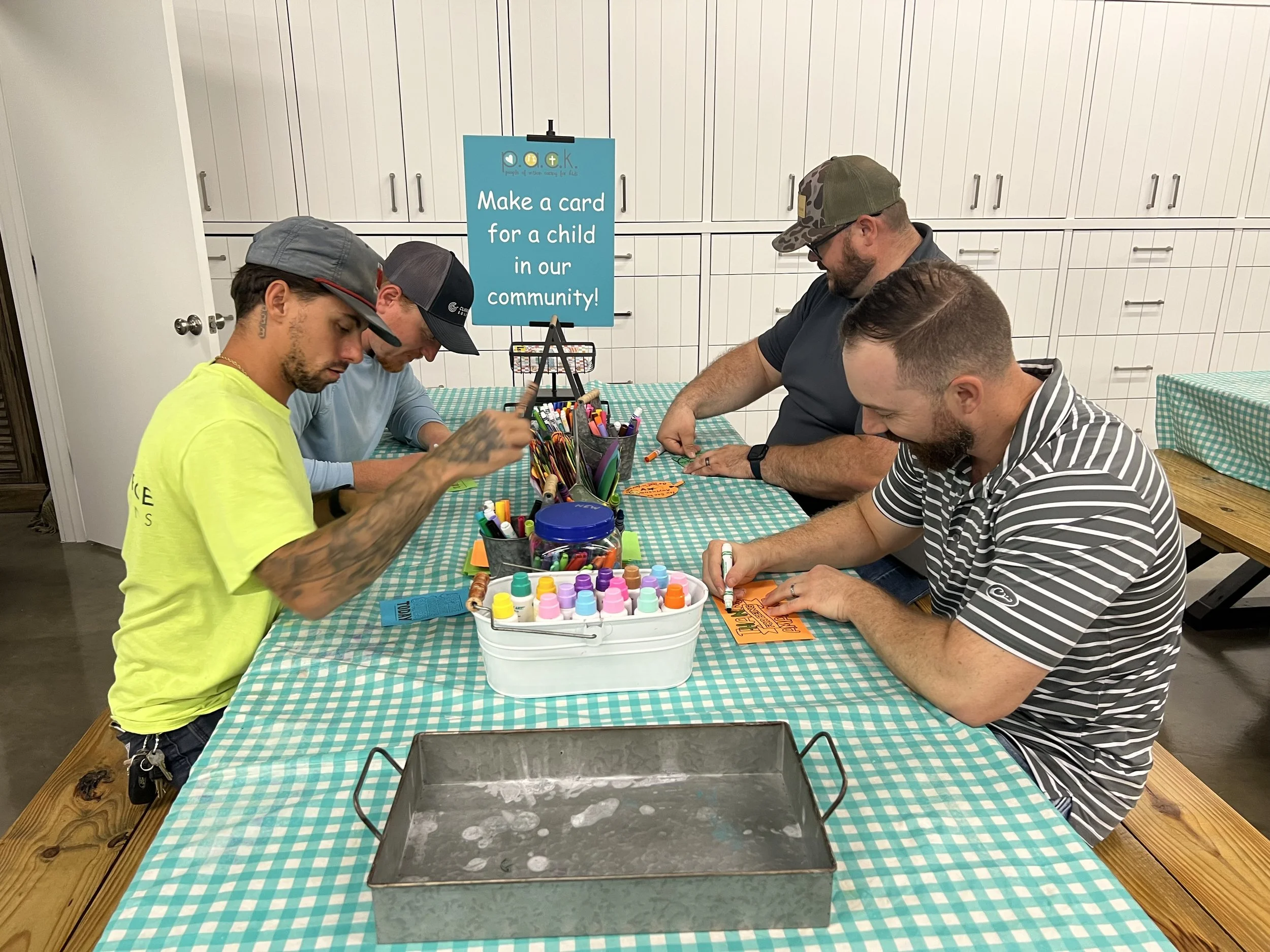 Four men at a table creating cards for children in the community for a volunteer event, with art supplies and a sign in the background.