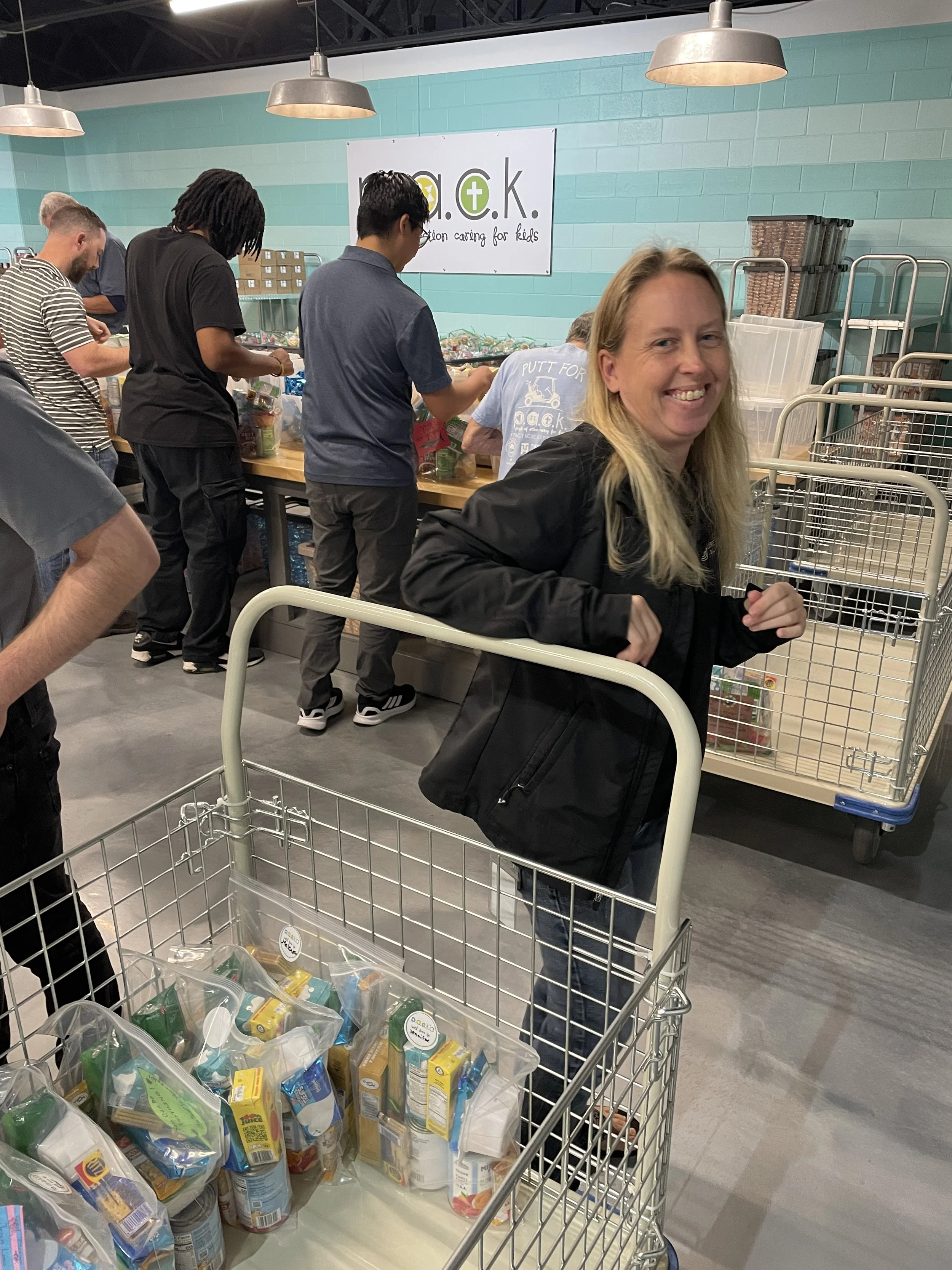 Smiling woman with blonde hair in a black jacket shopping at a food bank, standing behind a cart filled with packaged food, with people in the background volunteering and sorting donations.