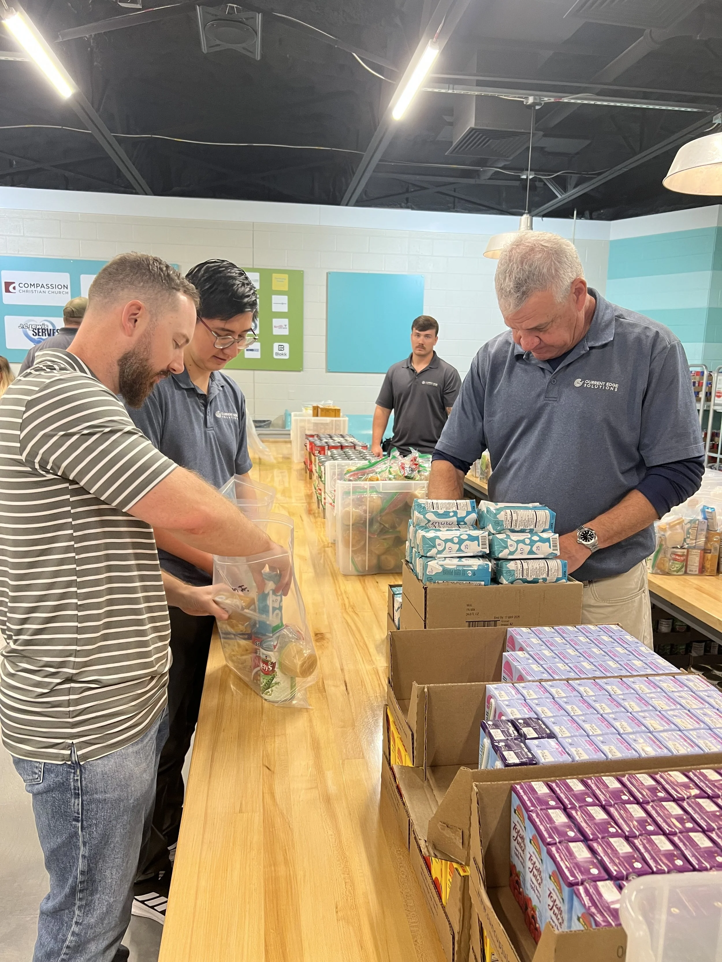 People packing food items at a food distribution event in a community center or church, with a variety of packaged goods on the table.