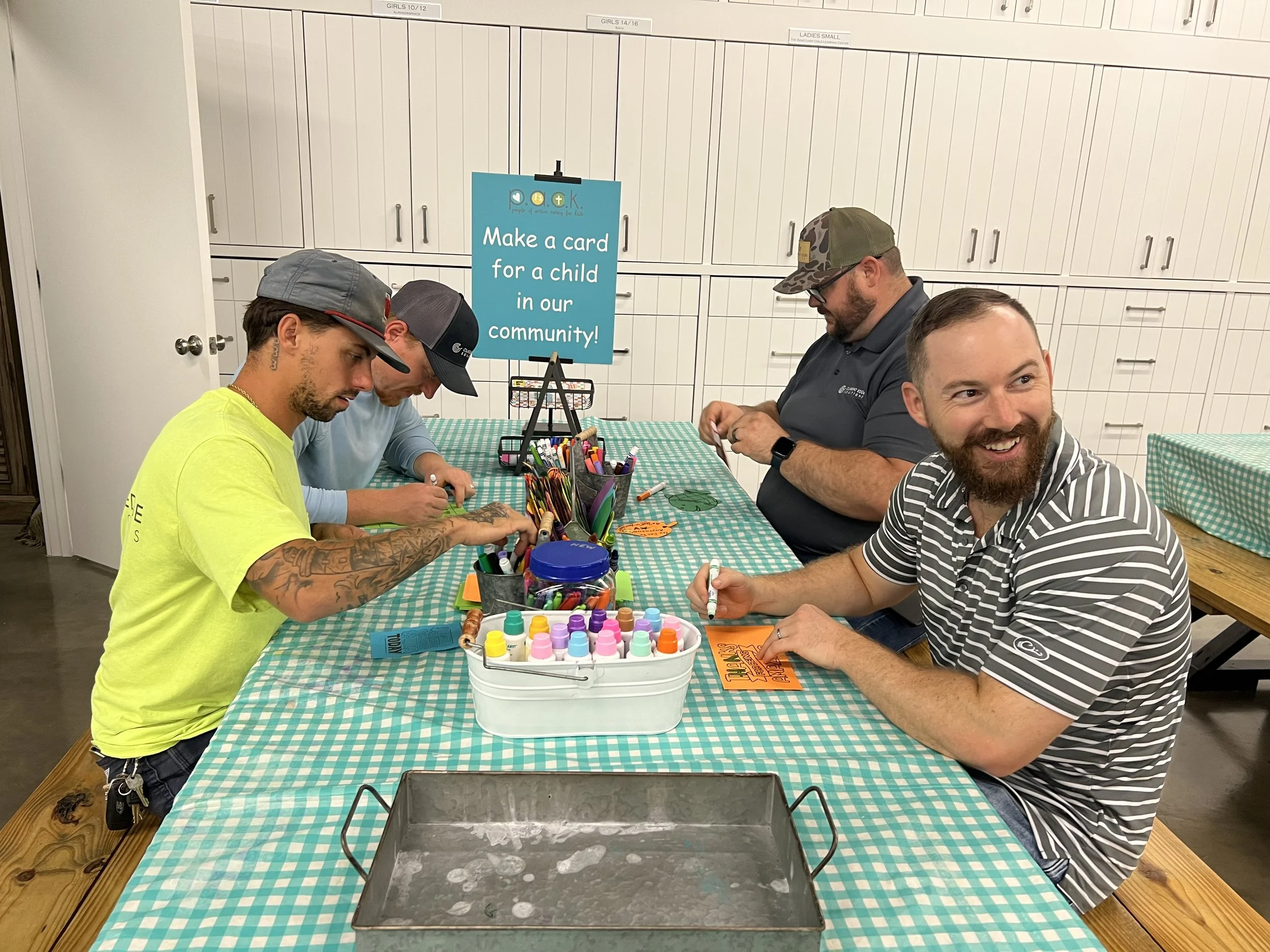 Four men sit at a long table covered with a turquoise checkered tablecloth, engaged in craft activity. In the foreground, a smiling man with a beard in a striped shirt holds a marker and cuts a card. Next to him, a man in a cap and sunglasses appears