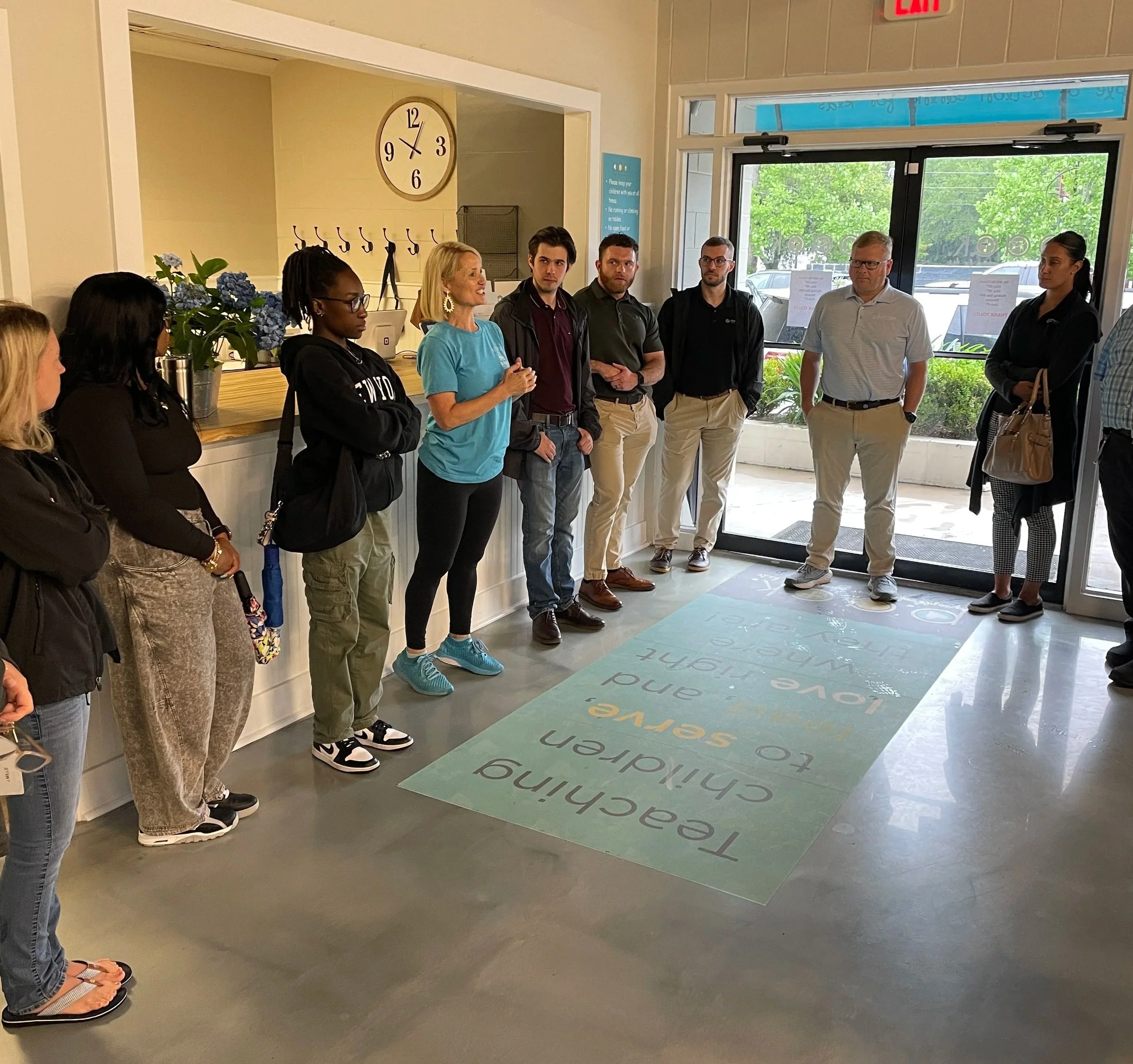 A group of people standing inside a hotel or office lobby near a large glass door, listening to a woman speak. There is a clock on the wall and a colorful mat on the floor with motivational words.