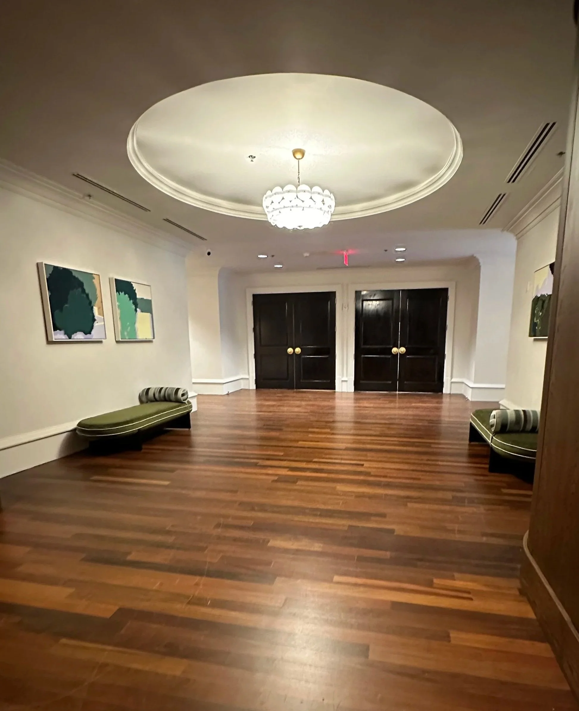 Empty hotel lobby with wooden floor, green upholstered benches, framed artwork on white walls, and a central chandelier.
