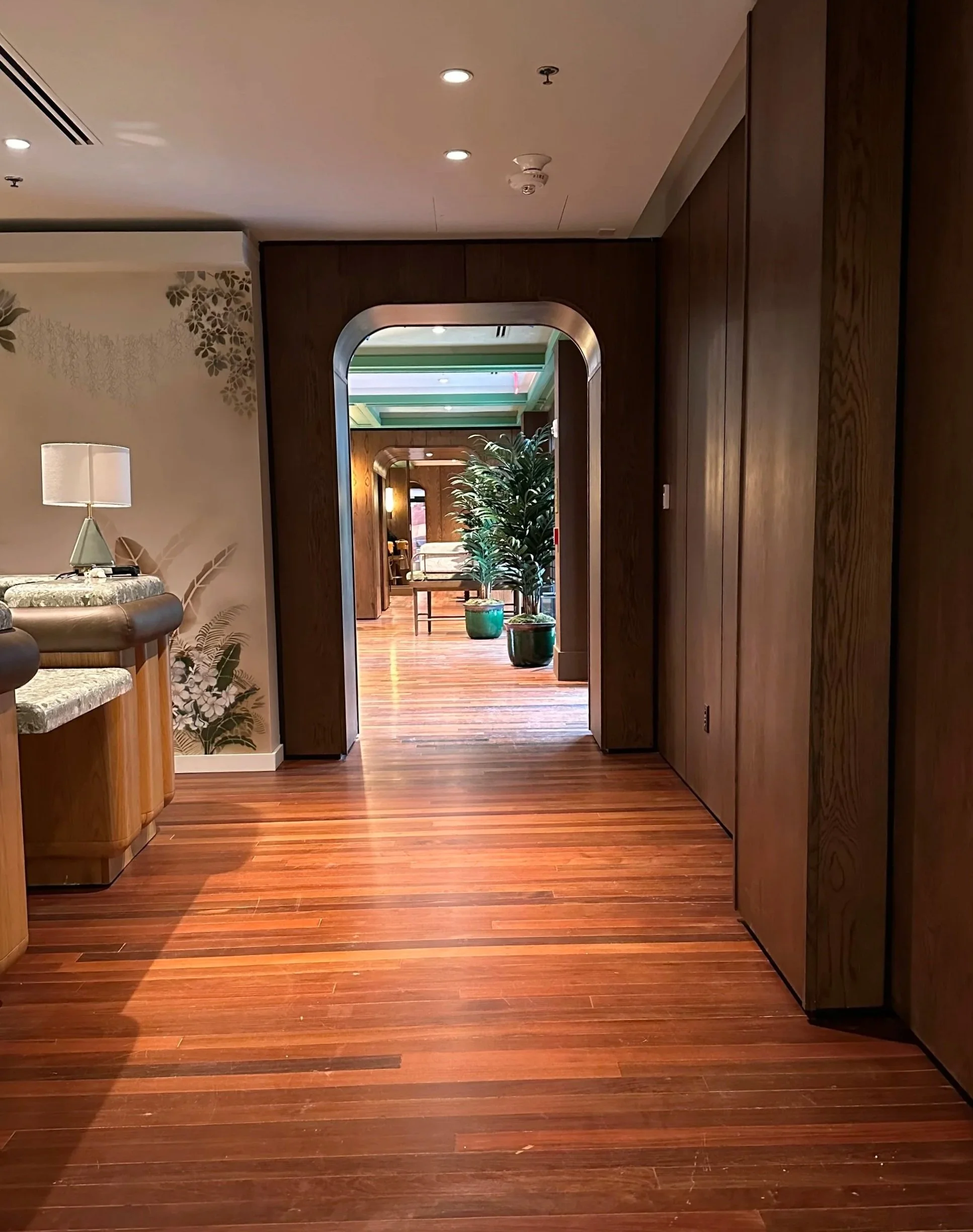 Indoor hallway with wooden floors, decorated with potted plants and furniture, leading to a larger room with seating.
