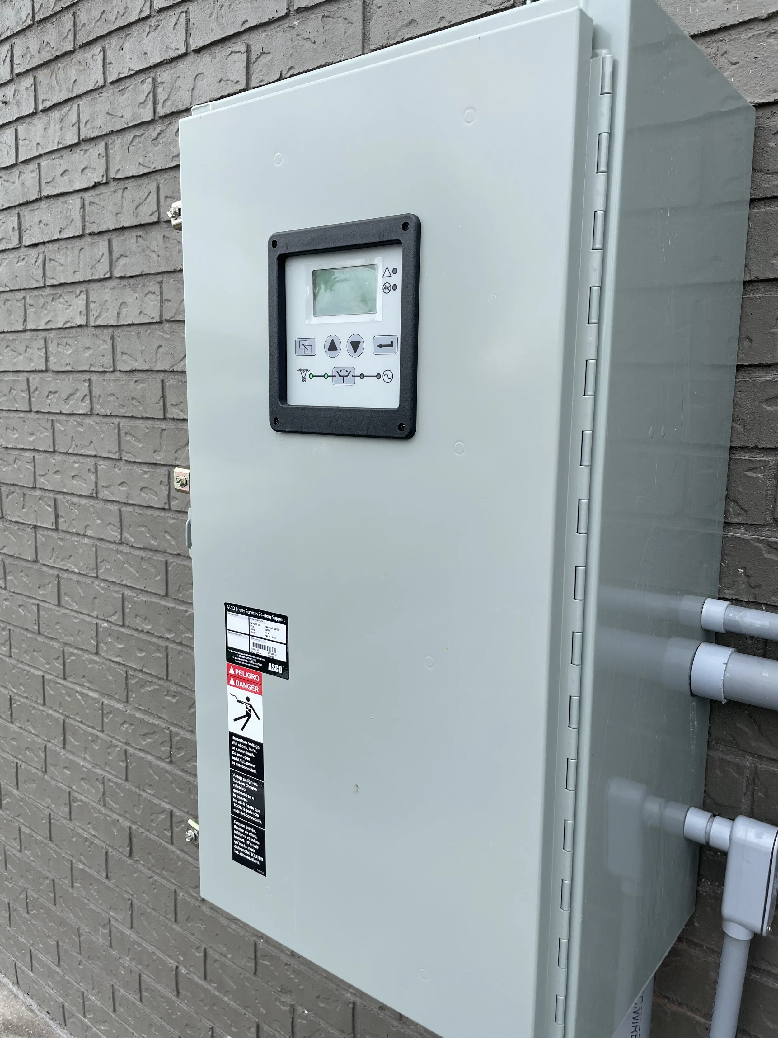 Electrical control box mounted on a brick wall with warning labels and a digital control panel.