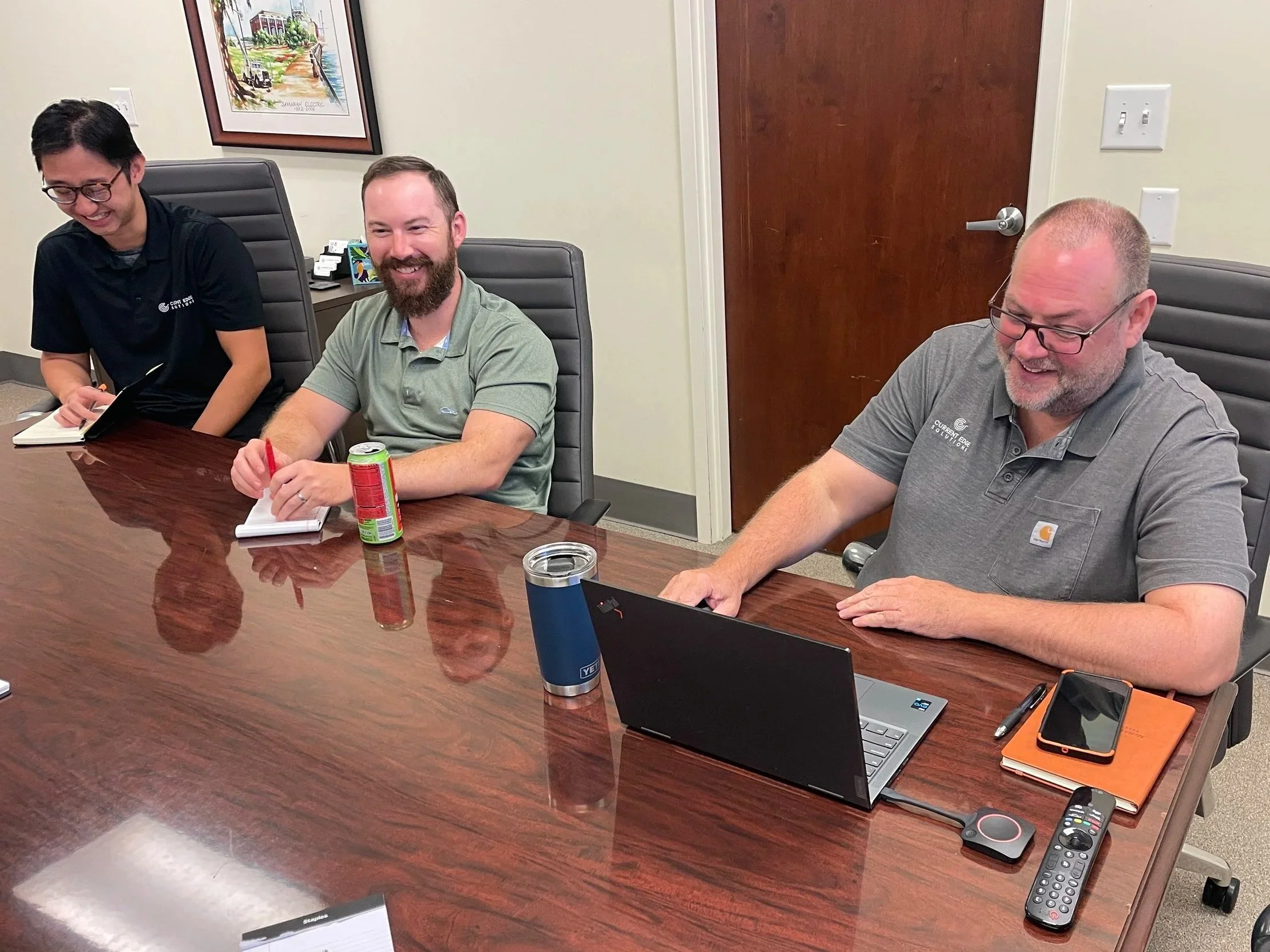 Three men sitting at a conference table, smiling. The man on the left is taking notes, the man in the middle is holding a red pen and has a can of soda in front of him, and the man on the right is using a laptop with a smartphone, a notebook, and a remote control nearby.