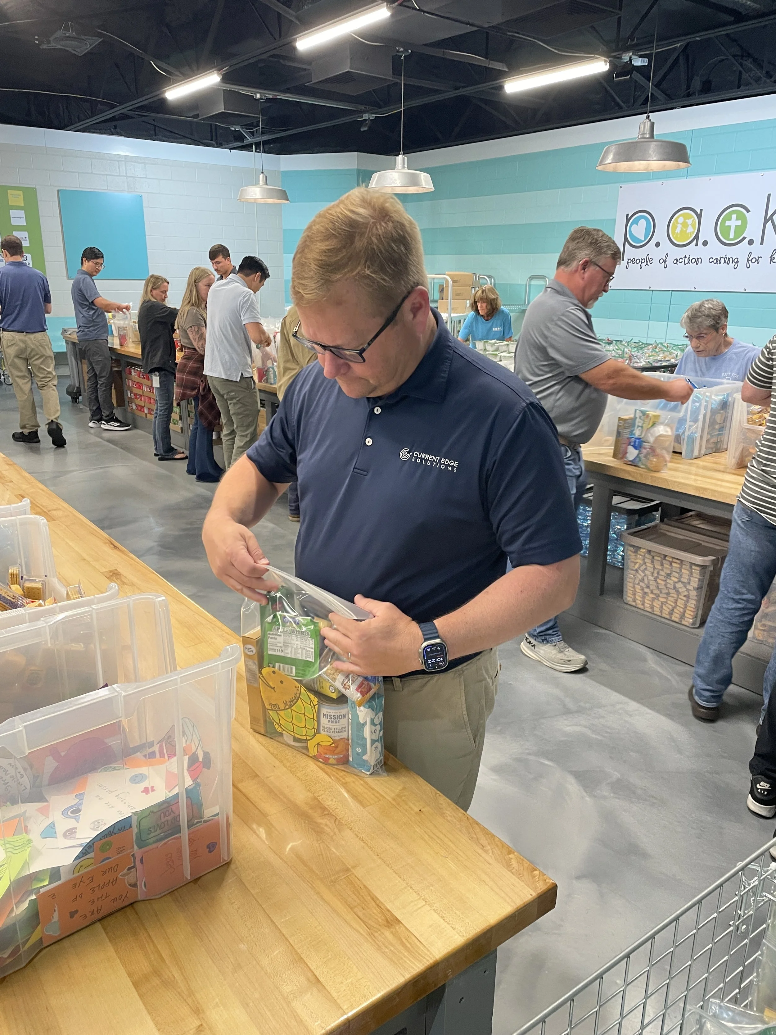 People volunteering at a food packaging event, filling bags with food items at a long table in a community center or warehouse.