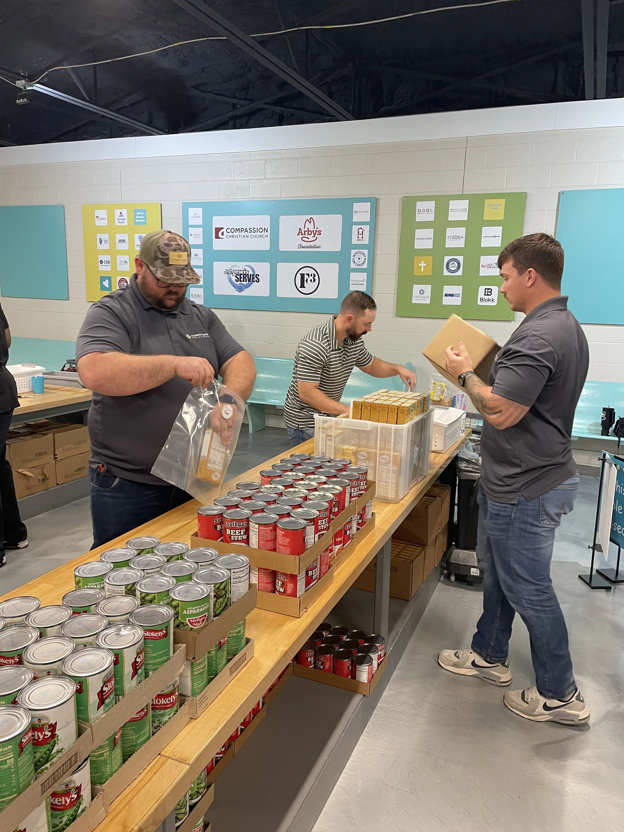 Three men organizing canned and boxed food items on a wooden table at a food pantry, with colorful business logos and church names displayed on the wall behind them.
